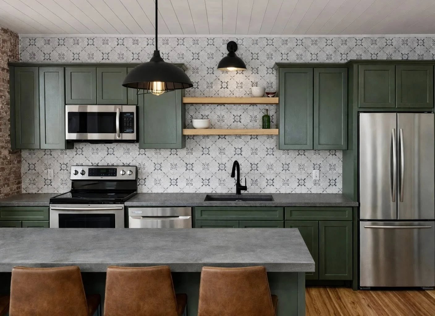 Modern kitchen with green cabinets, stainless steel appliances, open wooden shelves with dishes and a small plant, patterned tile backsplash, black faucet, gray countertop, wooden flooring, and brown chairs in the foreground.