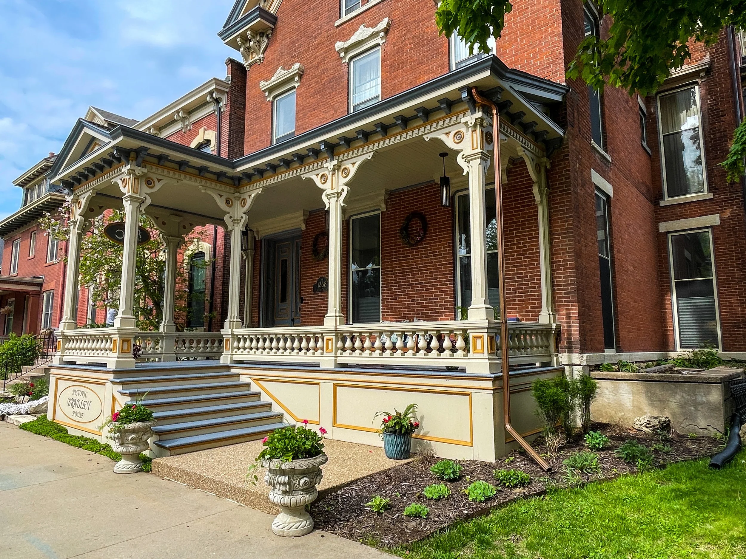 A Victorian-style brick house with decorative trim and a porch. The porch has ornate white columns, detailed woodwork, and a railing. There are plants and flowers in pots on the porch steps and around the house. The house has multiple windows and a sign that says 'Historic Bradford House' near the stairs. The scene is outdoors with a blue sky and trees nearby.