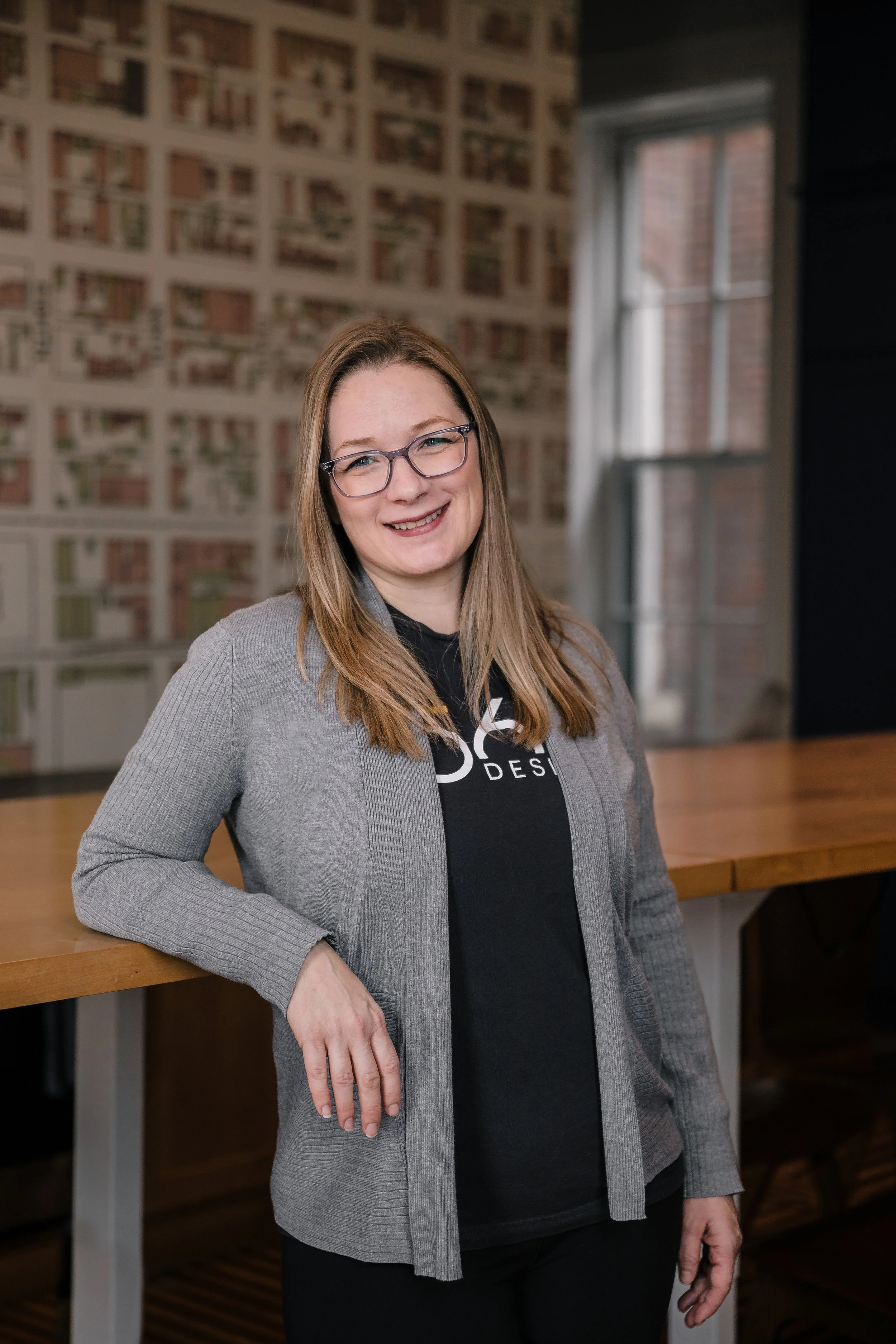 A woman with glasses and long hair, wearing a grey blazer over a black T-shirt, is smiling and standing in front of a brick wall with a large window visible in the background.