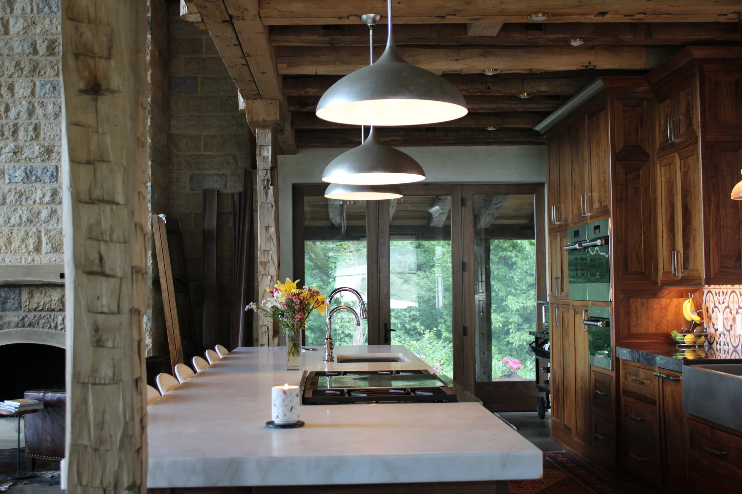 A rustic kitchen with wooden beams on the ceiling, pendant lights, a white kitchen island with a vase of flowers, and a large window overlooking greenery.