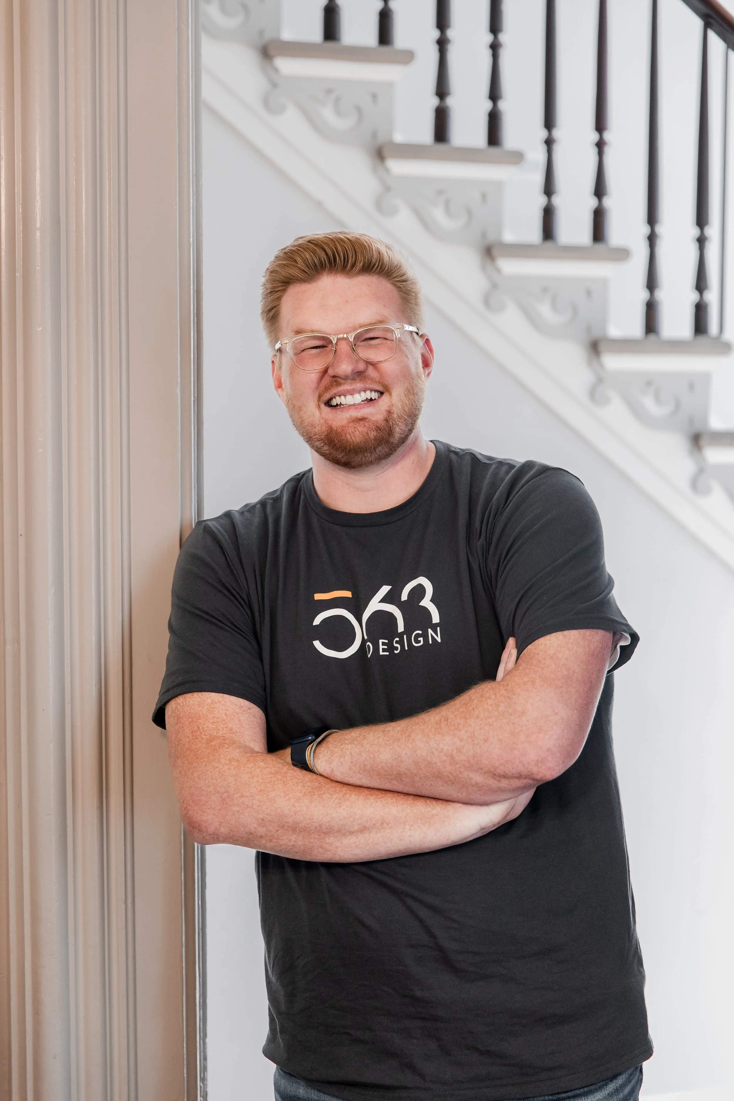 A smiling man with glasses and a beard standing with arms crossed against a beige wall and a staircase in the background.