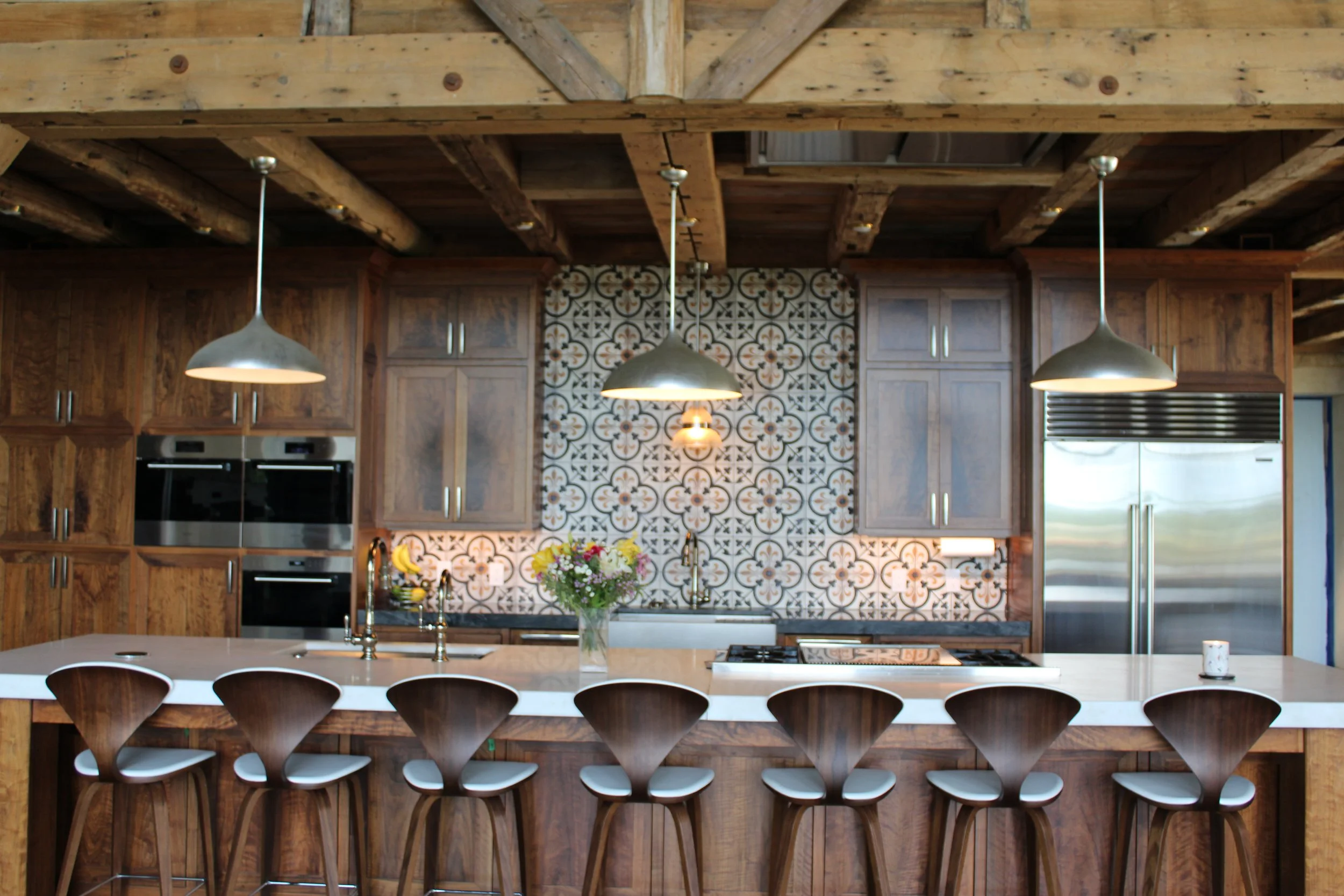 Modern kitchen with wooden cabinets, patterned tile backsplash, and three metal pendant lights hanging over a large island with bar stools.