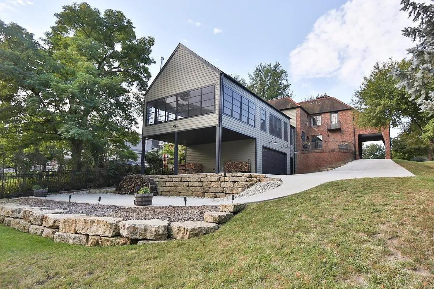 A modern house with a second-floor enclosed porch, supported by black columns, situated on a sloped lawn with stonework and a concrete pathway.