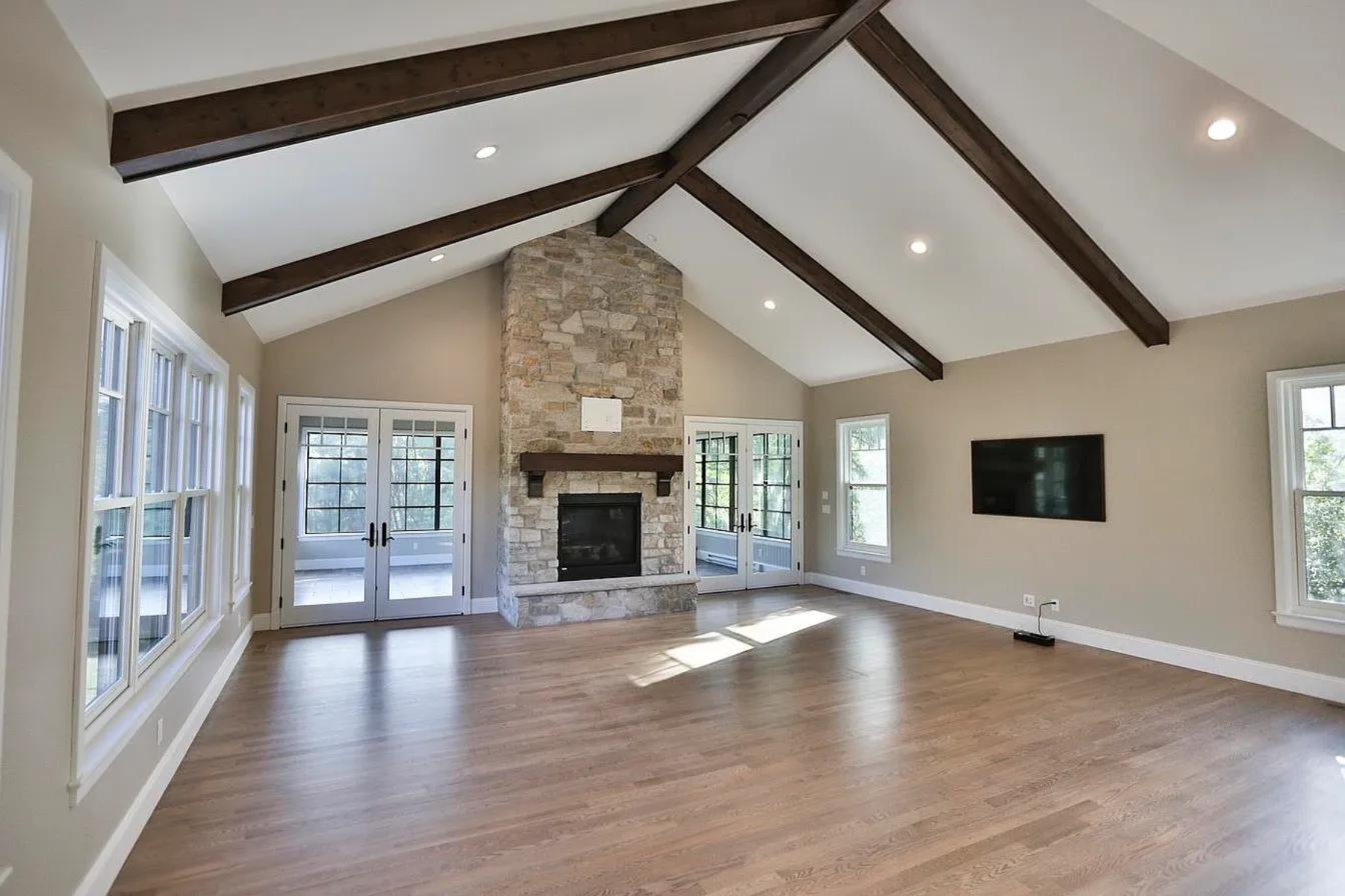 Living room with a stone fireplace, wooden ceiling beams, hardwood flooring, multiple large windows, and French doors leading outside, with a mounted flat-screen TV on the wall.