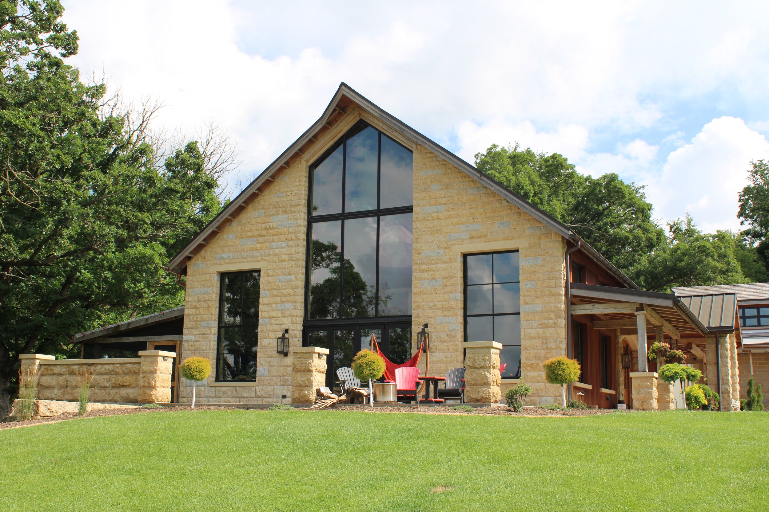 A modern house with large glass windows, stone exterior walls, and a sloped roof, surrounded by green trees and a well-maintained lawn.