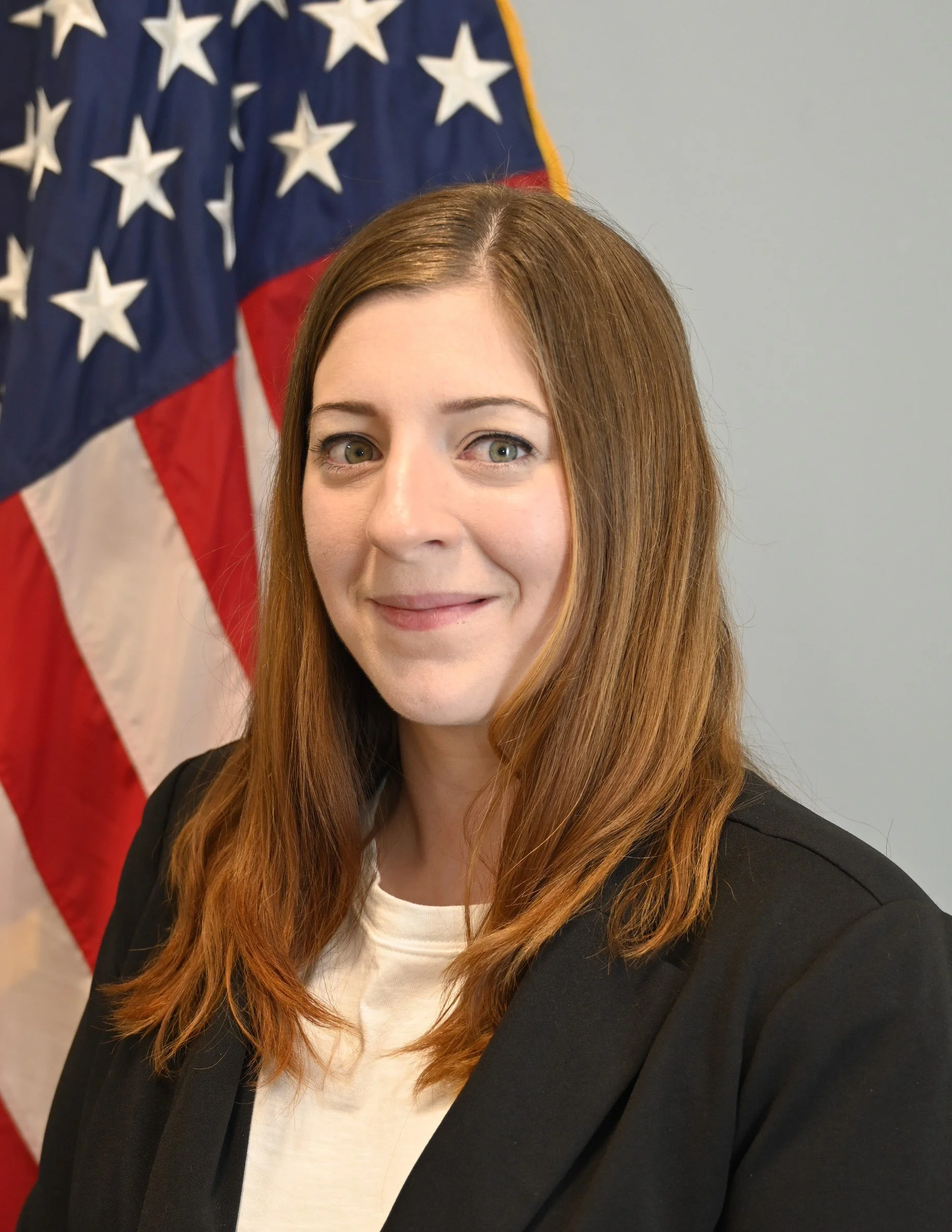 A woman with long red hair, wearing a black blazer and white shirt, standing in front of an American flag.
