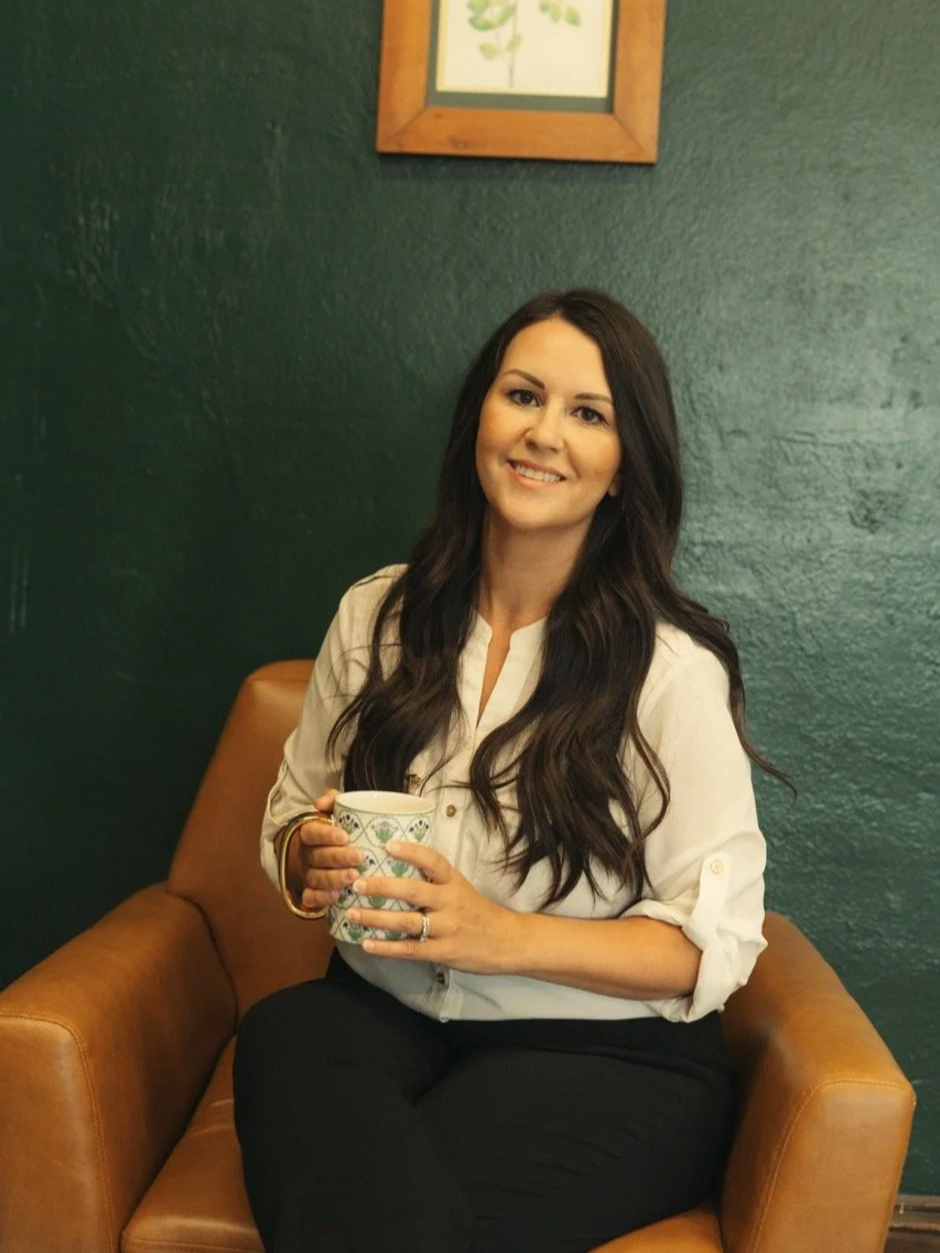 A woman with long dark hair sitting on a tan leather chair, smiling, holding a mug with a green and white pattern, in a room with dark green walls and a framed picture above her.