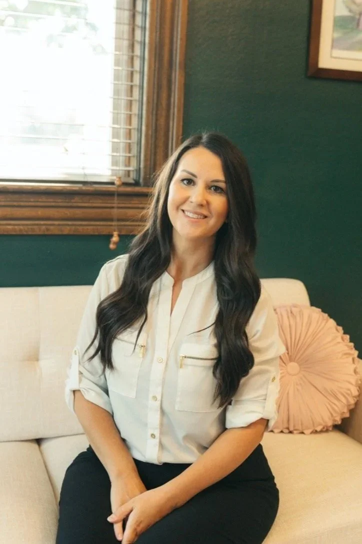 A woman with long dark hair, wearing a white shirt and black pants, sitting on a beige sofa in a cozy room with green walls and a window with blinds, smiling at the camera.