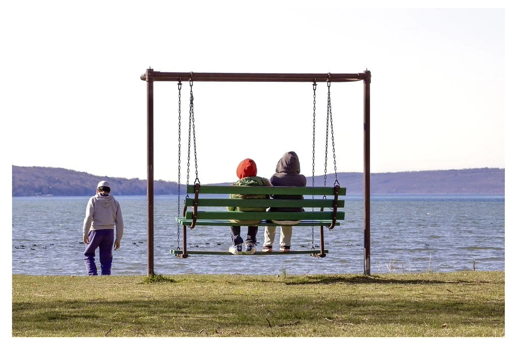 On a spring afternoon, two children sit on a swing bench overlooking Cayuga Lake while another explores the shoreline at Stewart Park in Ithaca, New York. 