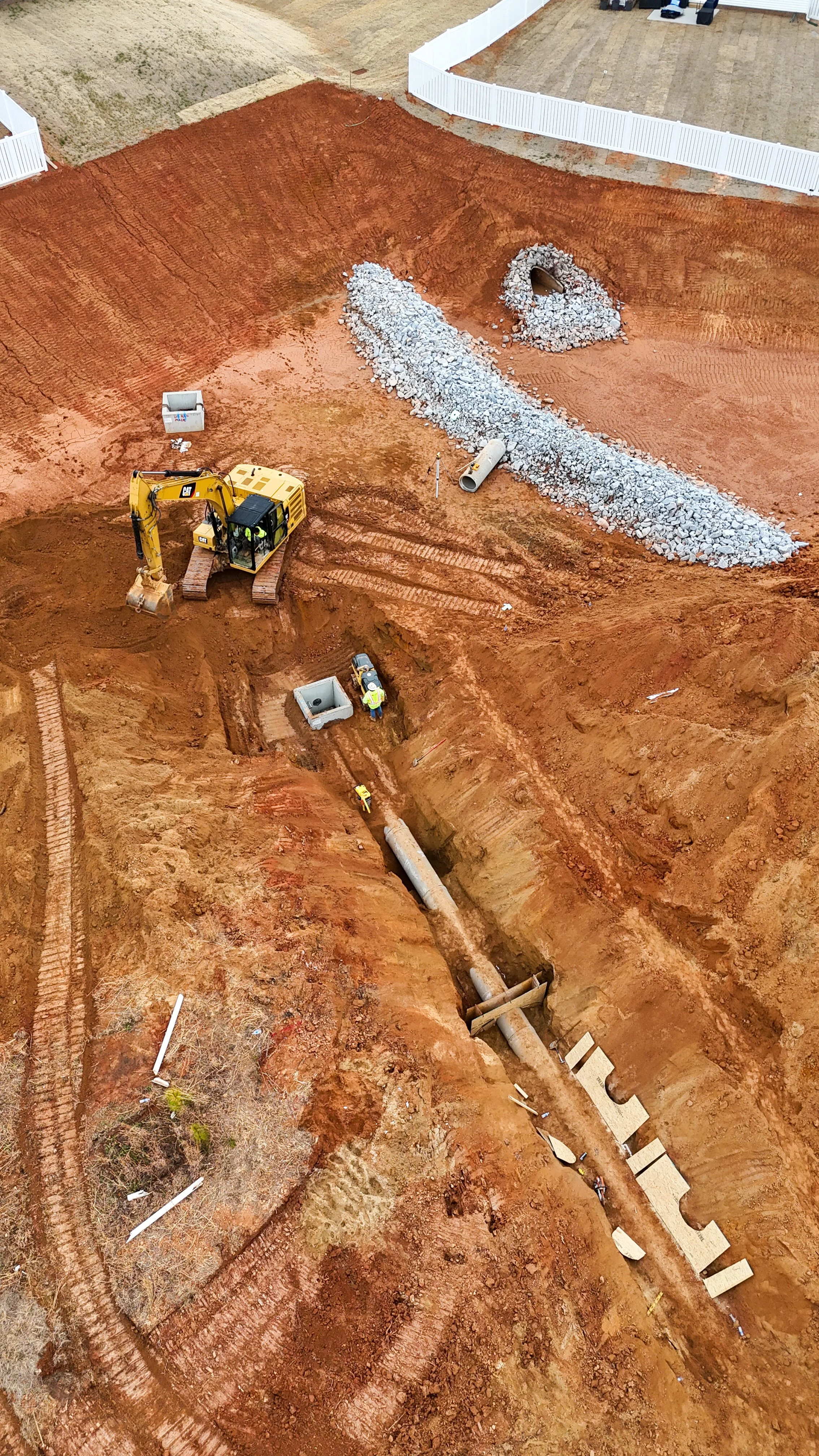 An aerial view of a construction site showing excavation, large pipes, a yellow excavator, gravel pile, and workers.