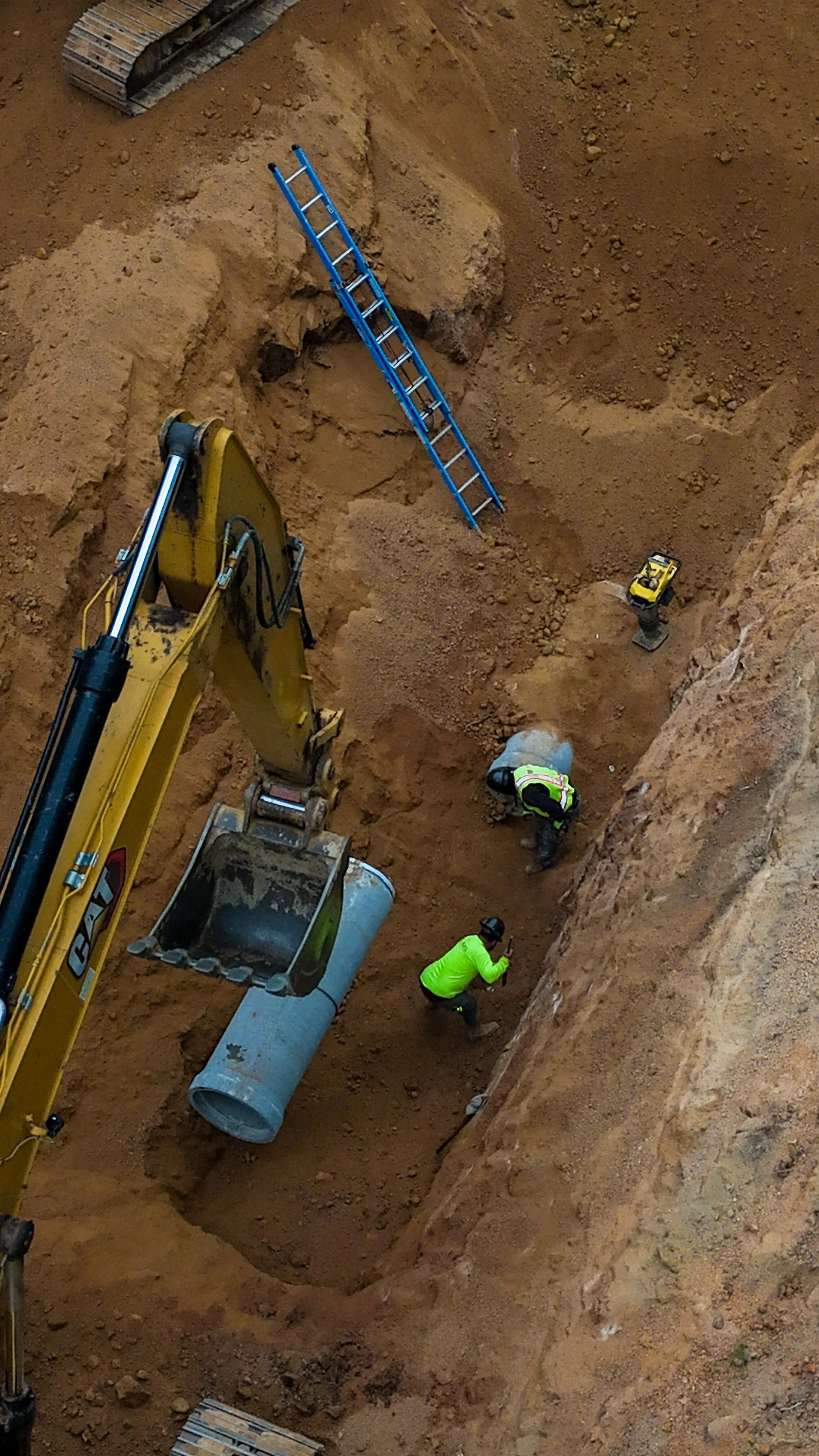 Construction workers in a trench installing large pipes with a yellow excavator and ladder nearby.