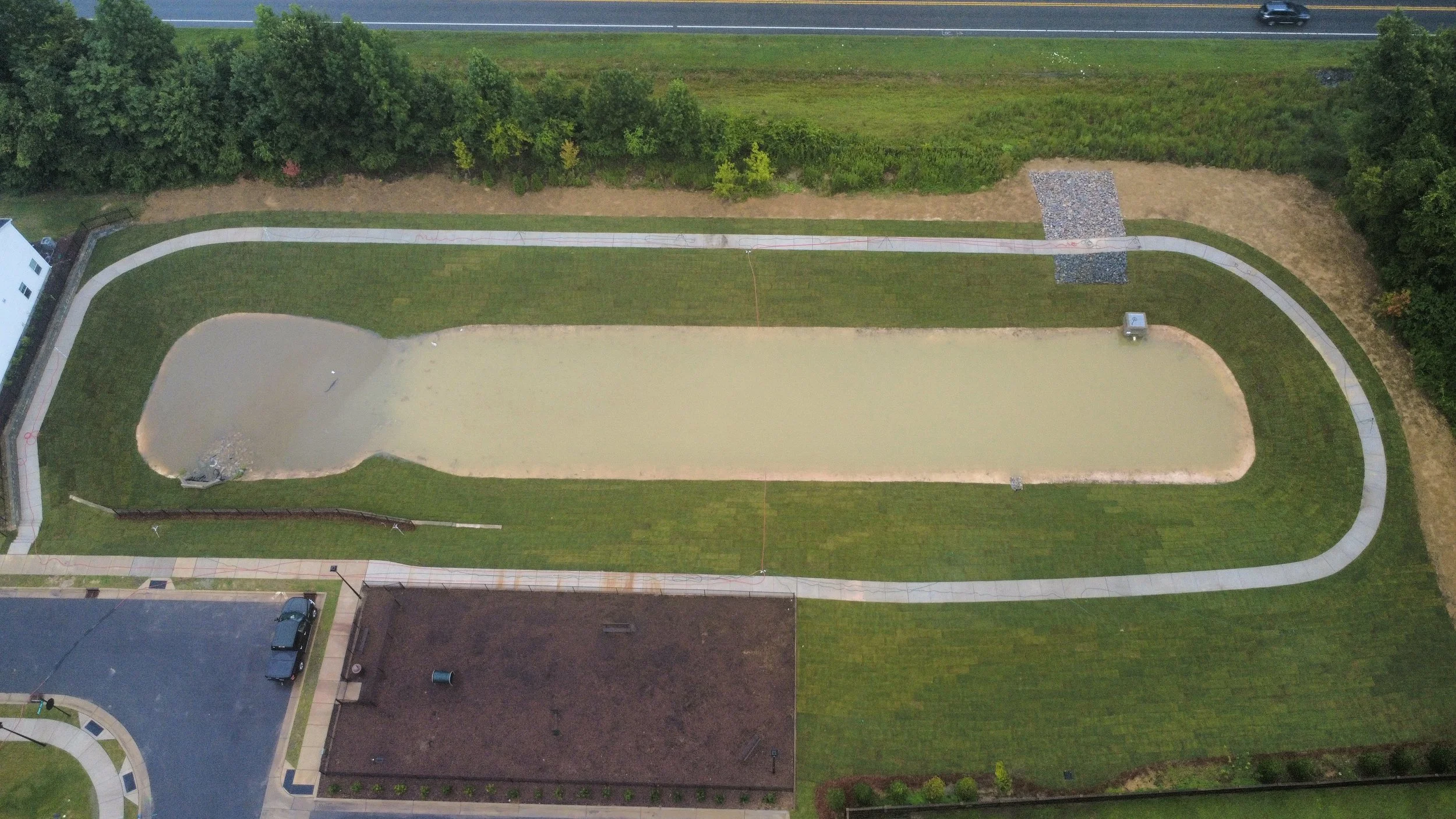 An aerial view of a backyard with a small pond, a swimming pool, a concrete pathway, and surrounding greenery, with a road and cars visible at the top of the image.