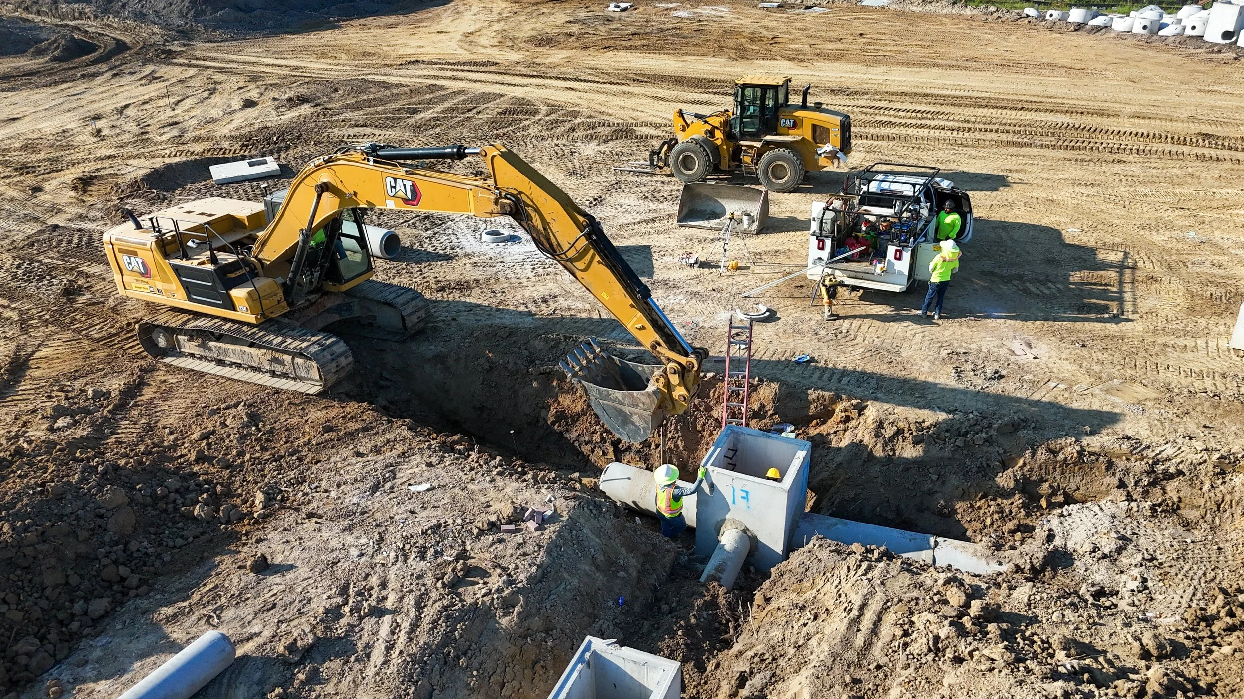 Construction site with two large yellow Caterpillar excavators and workers with safety gear. One worker is near a white utility truck, and the other appears to be near underground piping. The scene features dirt, construction equipment, and materials.