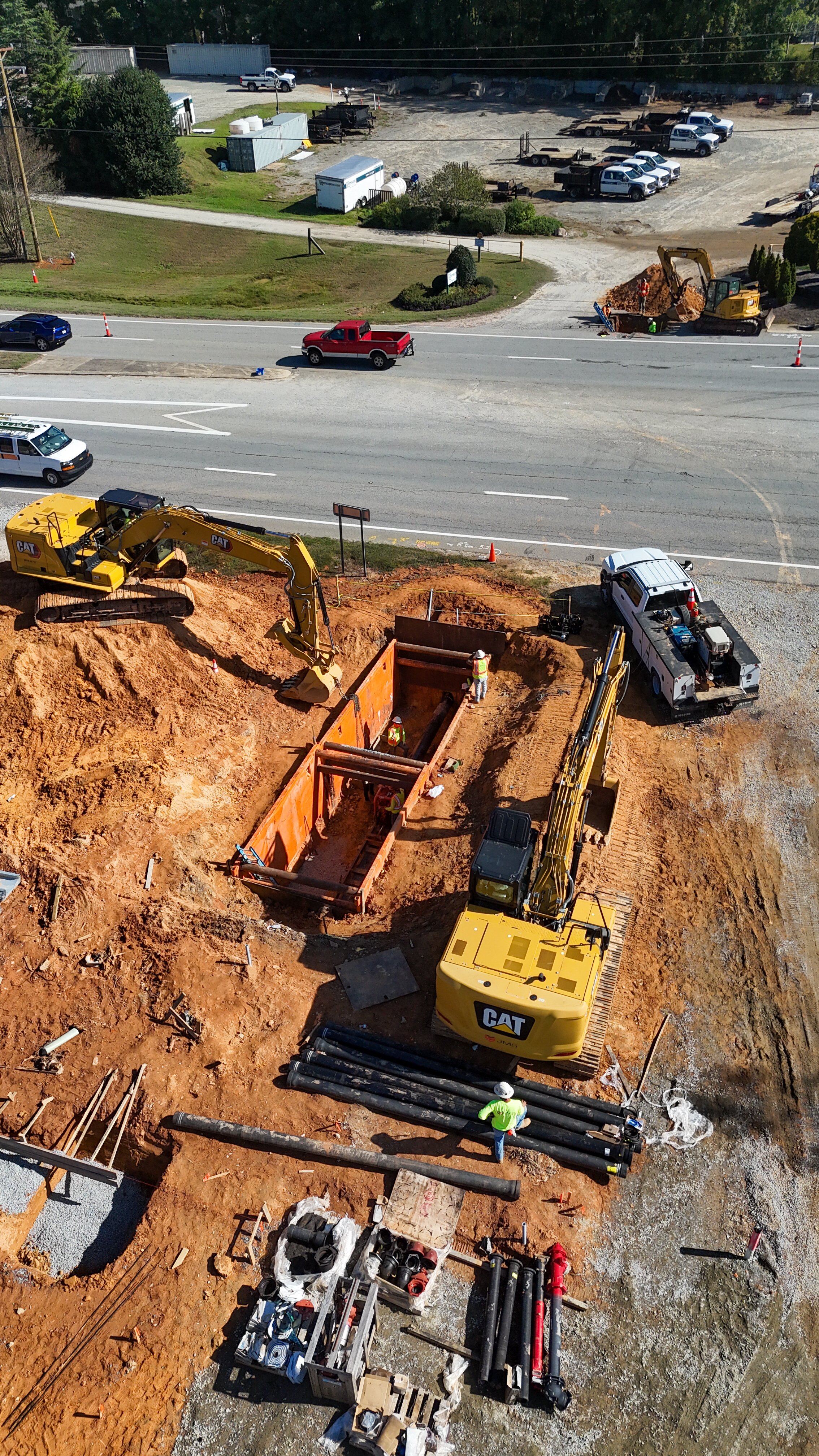 Construction site with excavators, workers, and piping work, next to a road with parked cars and trucks.