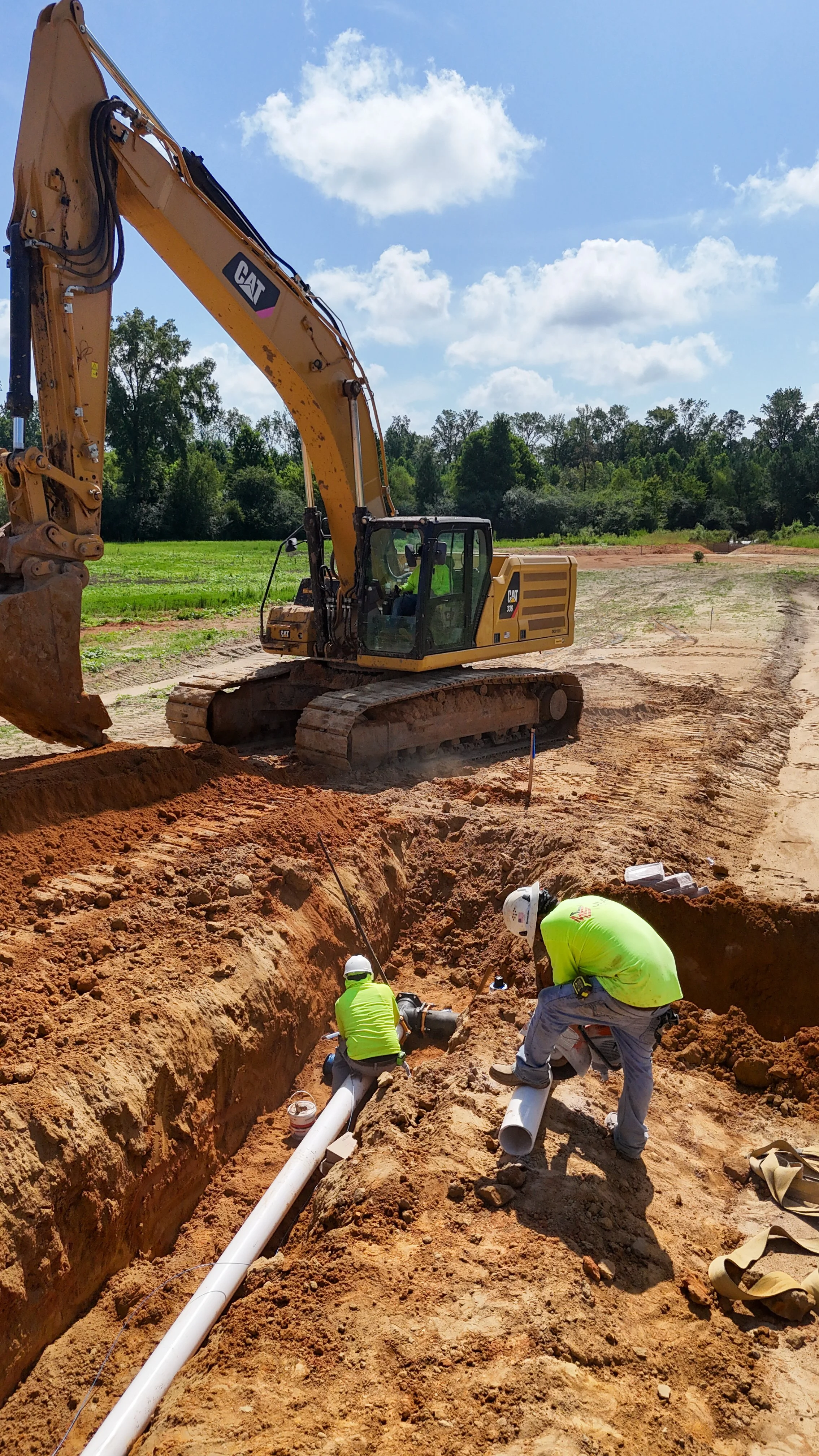 Construction workers and a large excavator working on underground piping on a construction site with green fields and trees in the background under a blue sky with clouds.