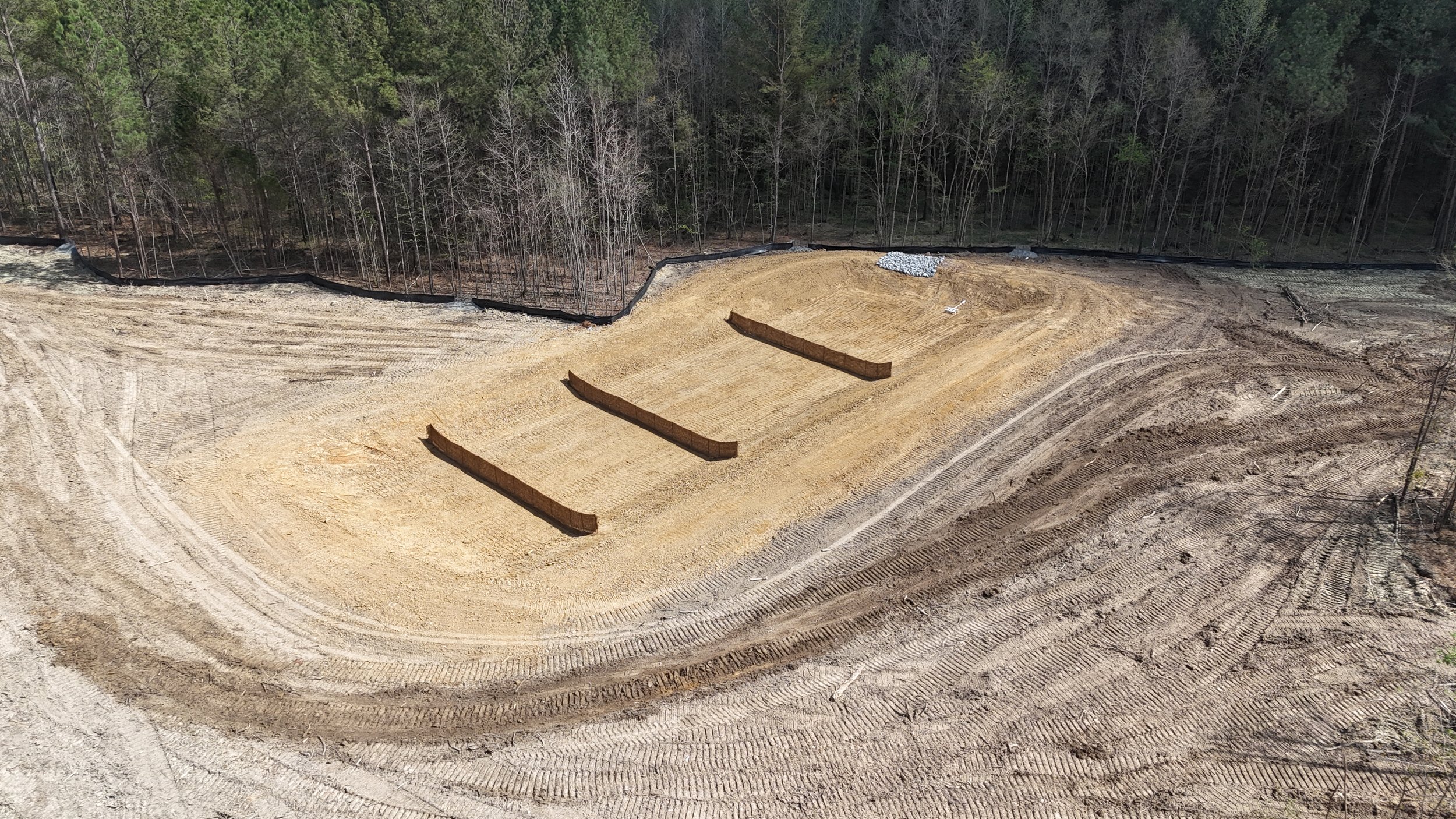 Construction site with three wooden barriers in a dirt clearing, surrounded by a black plastic barrier and a dense forest.