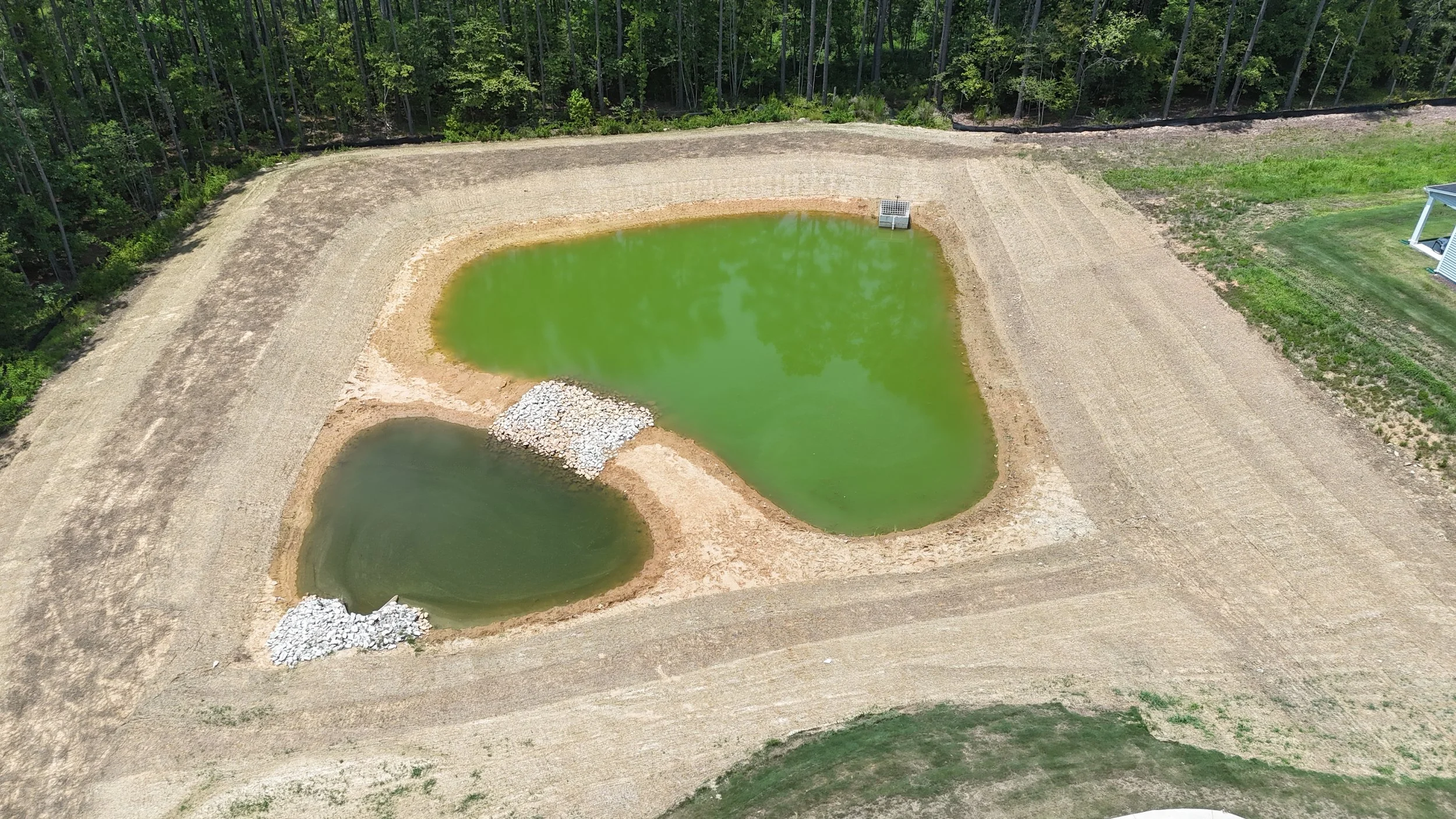 Aerial view of two small greenish ponds on a construction site, surrounded by dirt and trees.