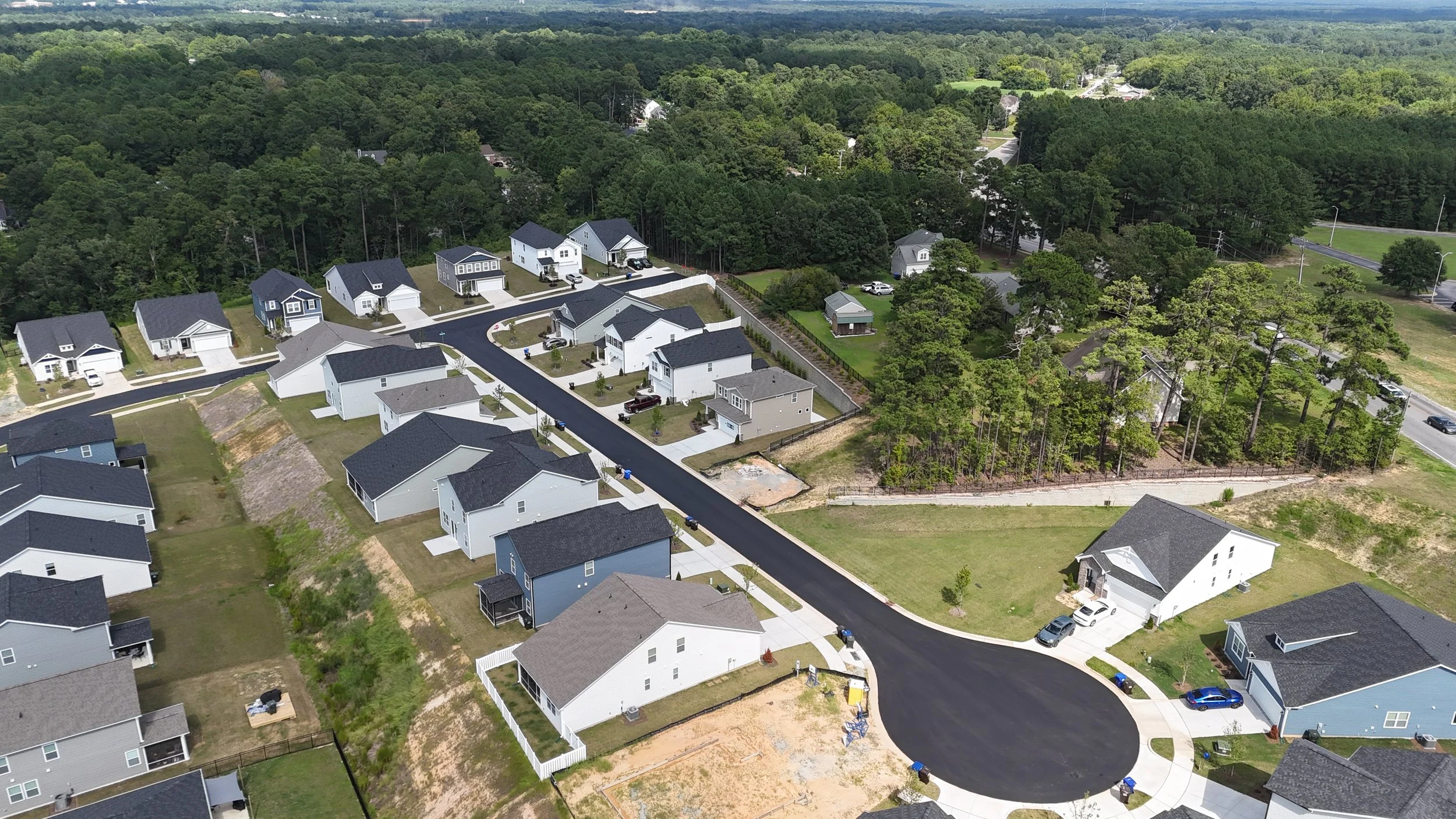 Aerial view of a suburban neighborhood with newly built houses, some with black roofs, surrounded by trees and greenery.
