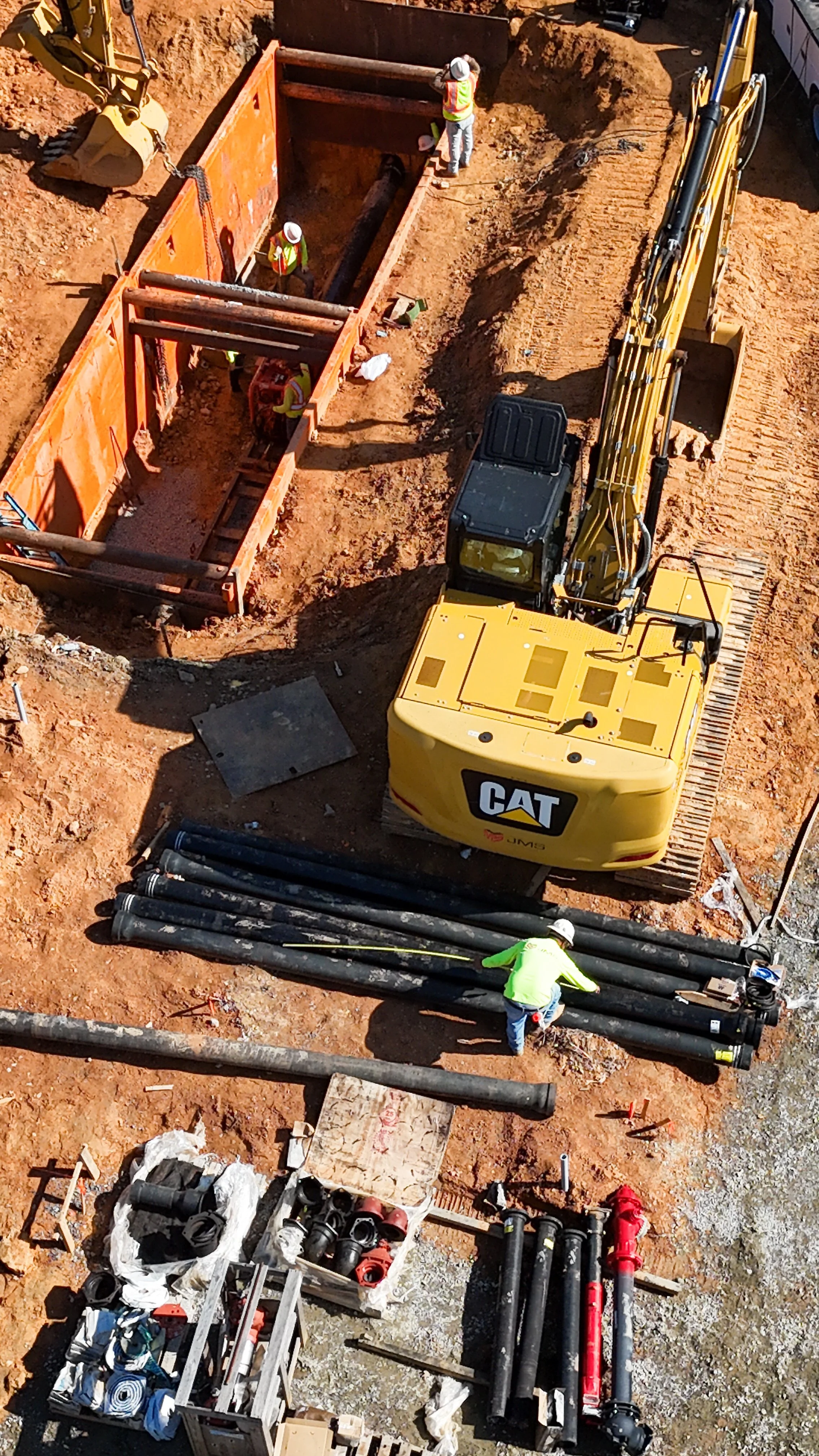 Construction workers are working on underground pipe installation. Equipment includes a large yellow CAT excavator, orange trench boxes, and various pipes and fittings on the ground.
