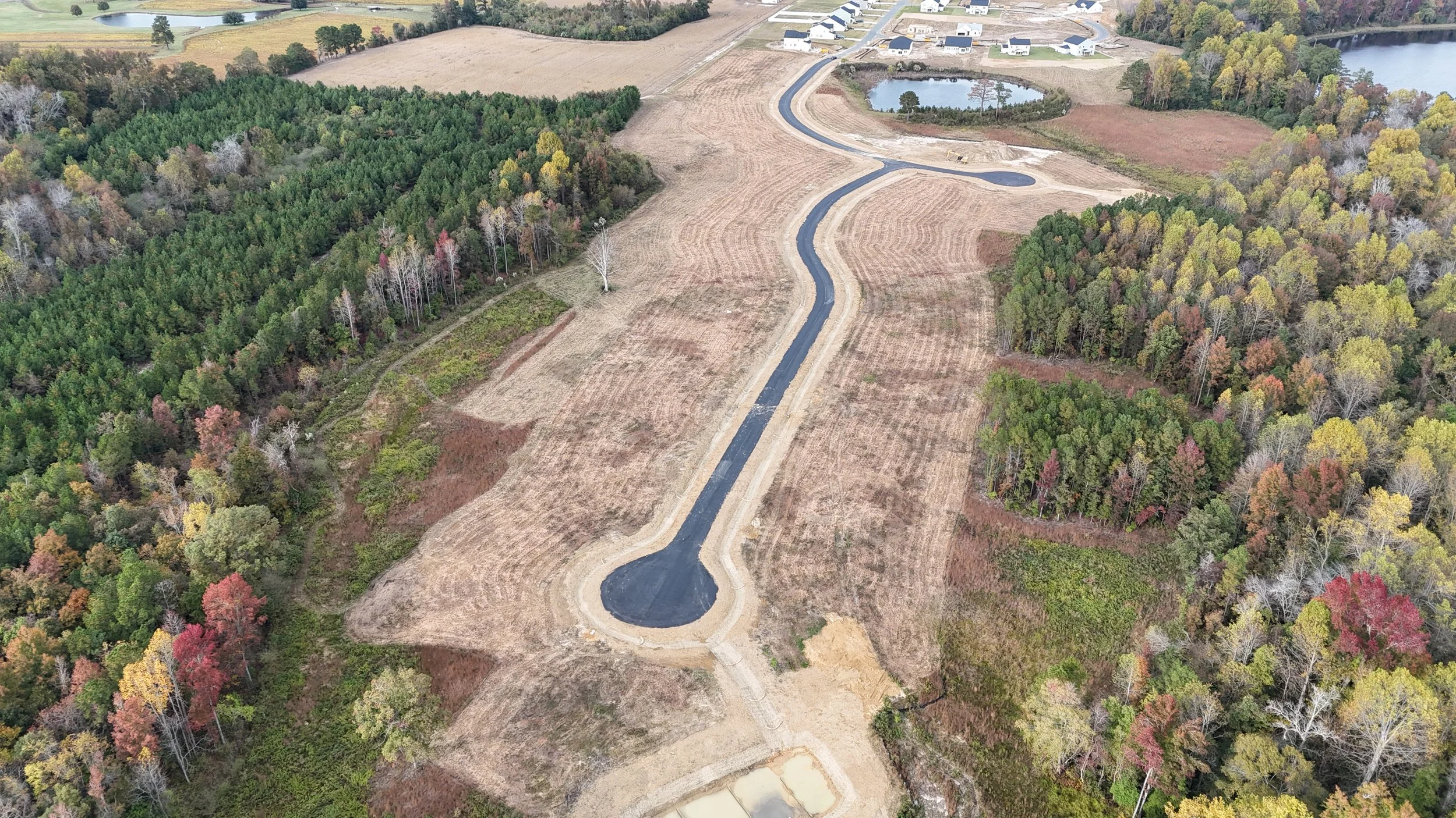 Aerial view of a construction site with a winding black asphalt road in the middle, surrounded by cleared land and dense forest with trees in fall colors, and nearby ponds and houses.