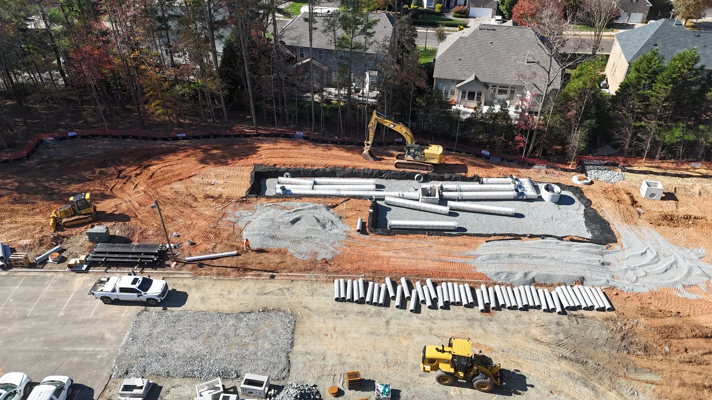 Construction site with large pipes, heavy machinery, and gravel, surrounded by trees and residential houses in the background.