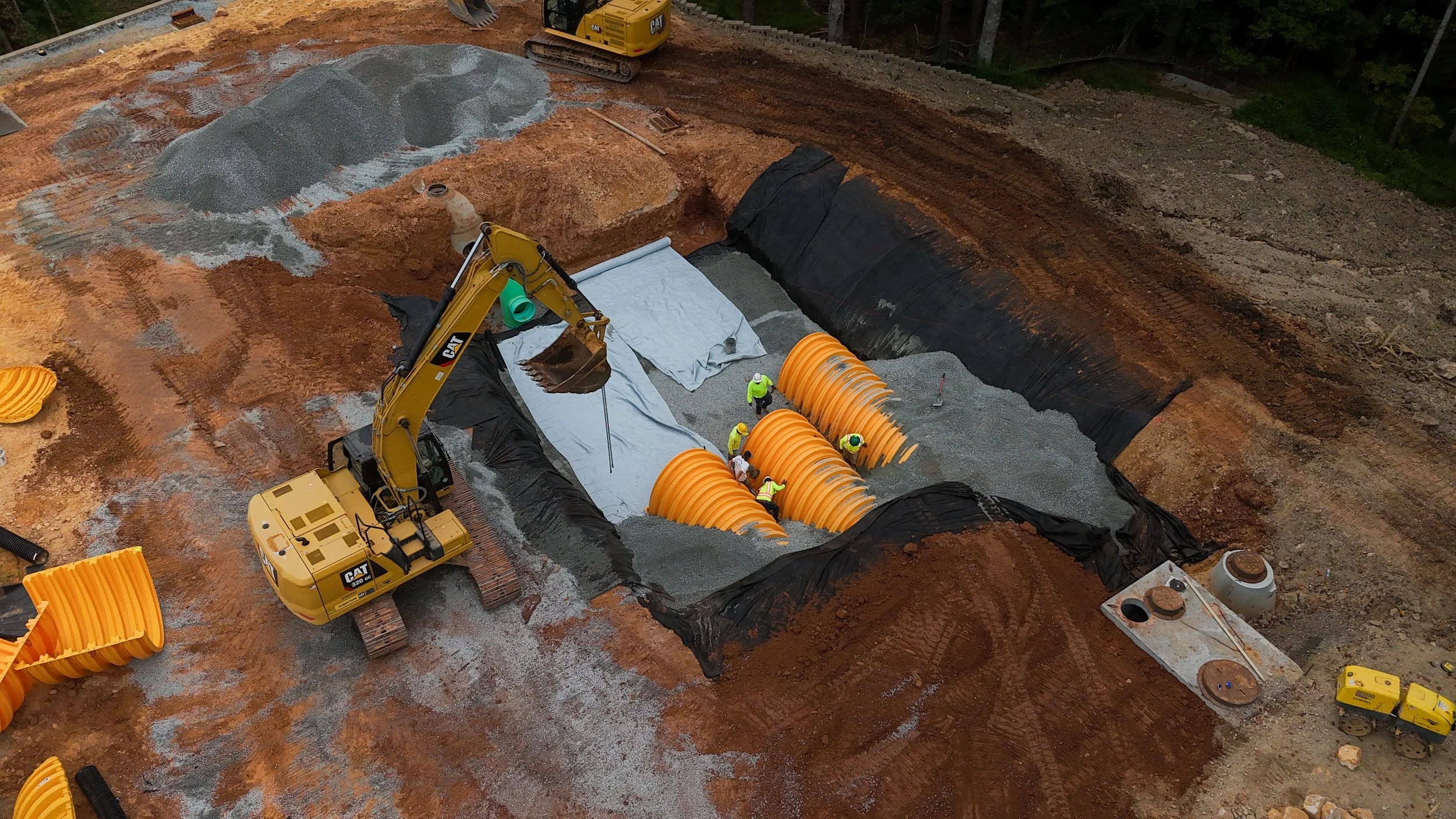 Construction site with workers laying foundation. Excavator, orange pipe, gravel, and black liner are visible.