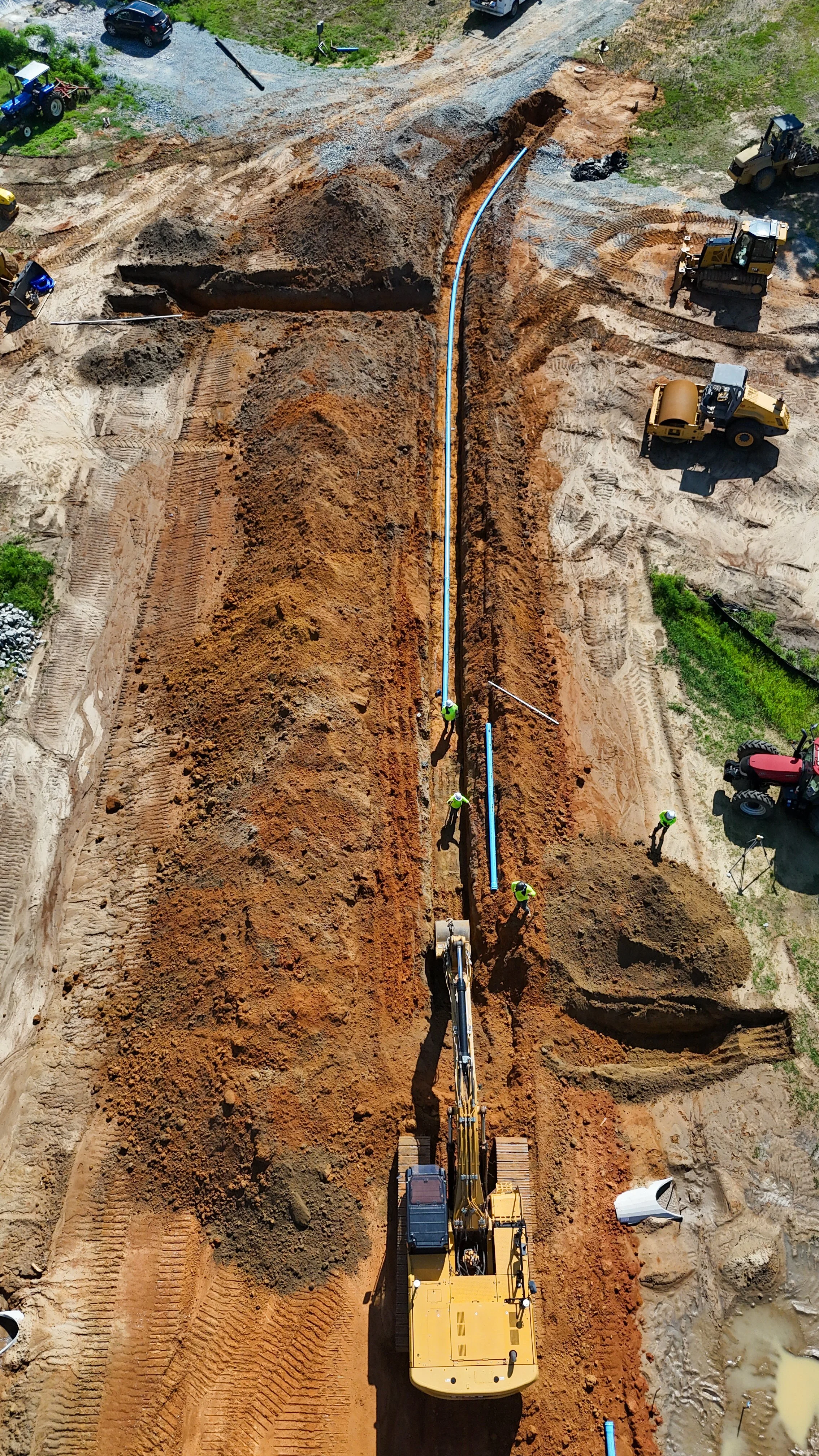 Construction site with excavator laying large blue pipes in a trench. Several workers in high-visibility vests are working nearby, and construction vehicles are present around the site.
