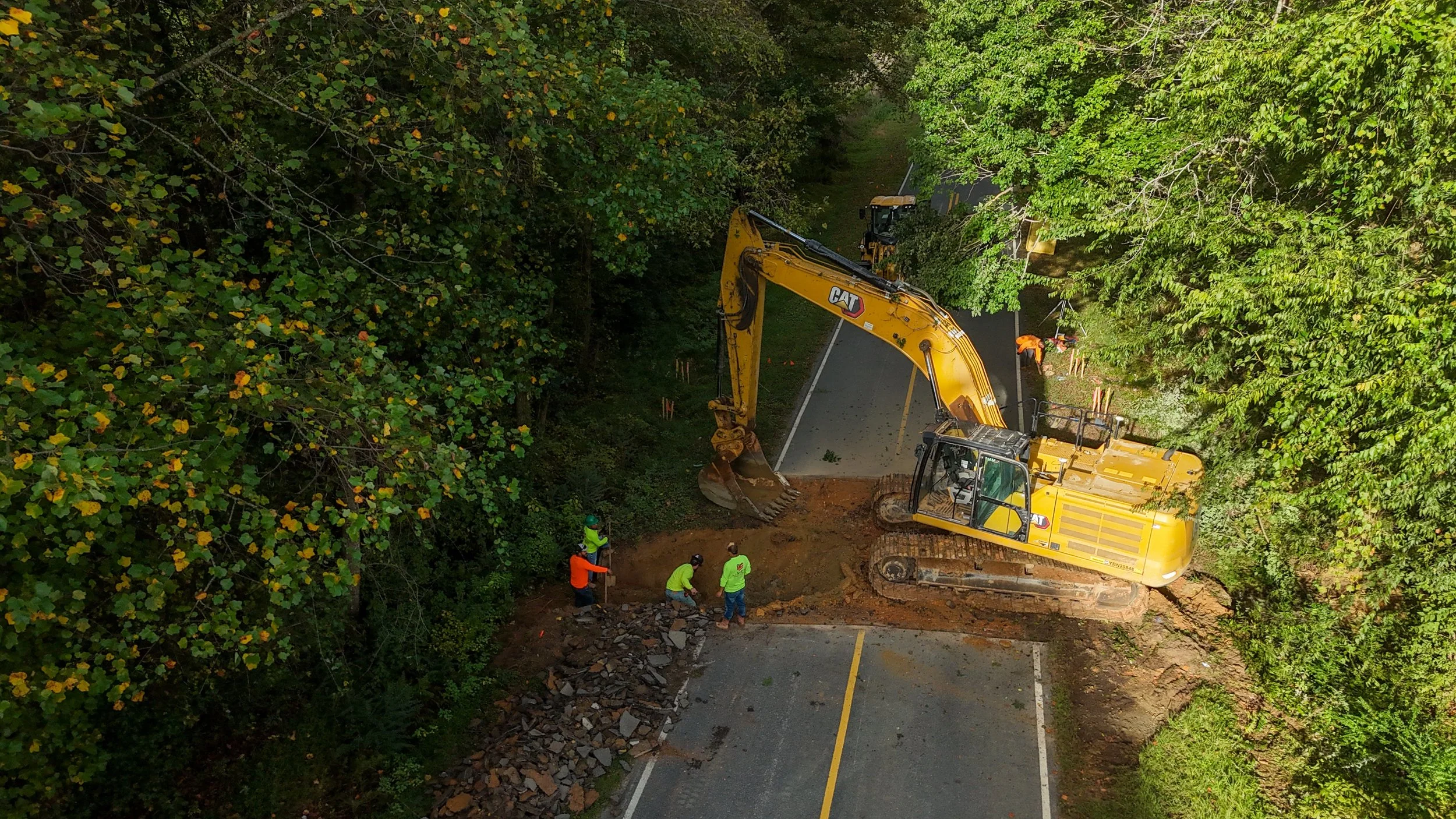 Construction crews working on road repair using an excavator in a wooded area.