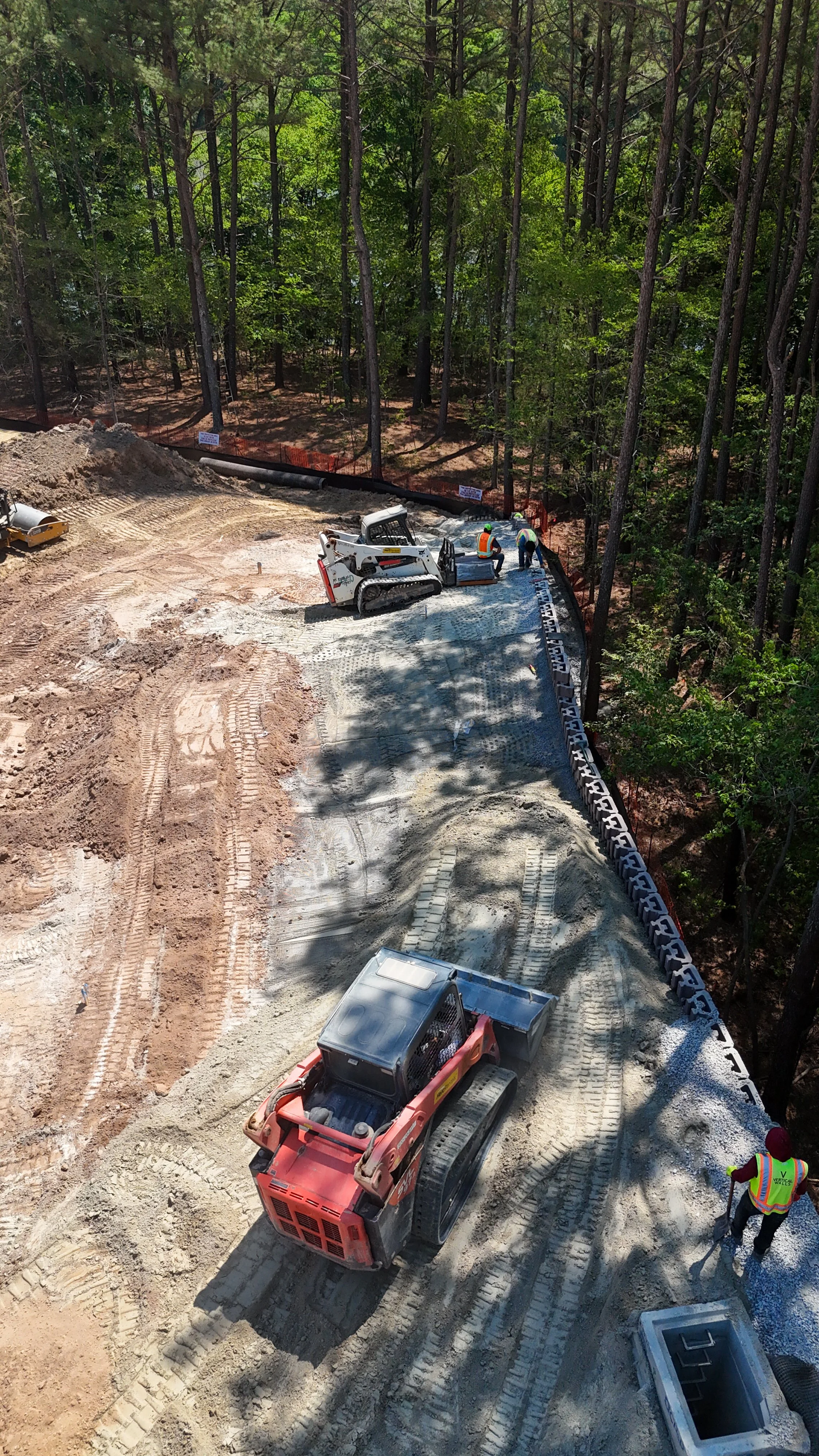Construction workers laying down a foundation with gravel, surrounded by trees, with machinery and construction supplies on site.