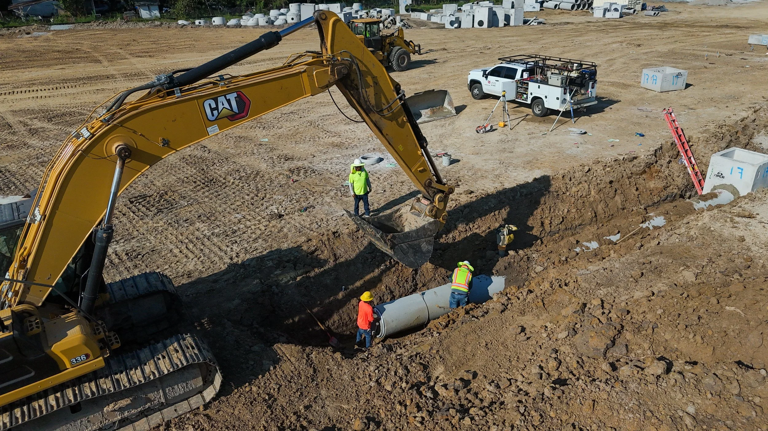 Construction site with a large yellow excavator digging a trench, workers installing large concrete pipes, and various construction vehicles and equipment in the background.