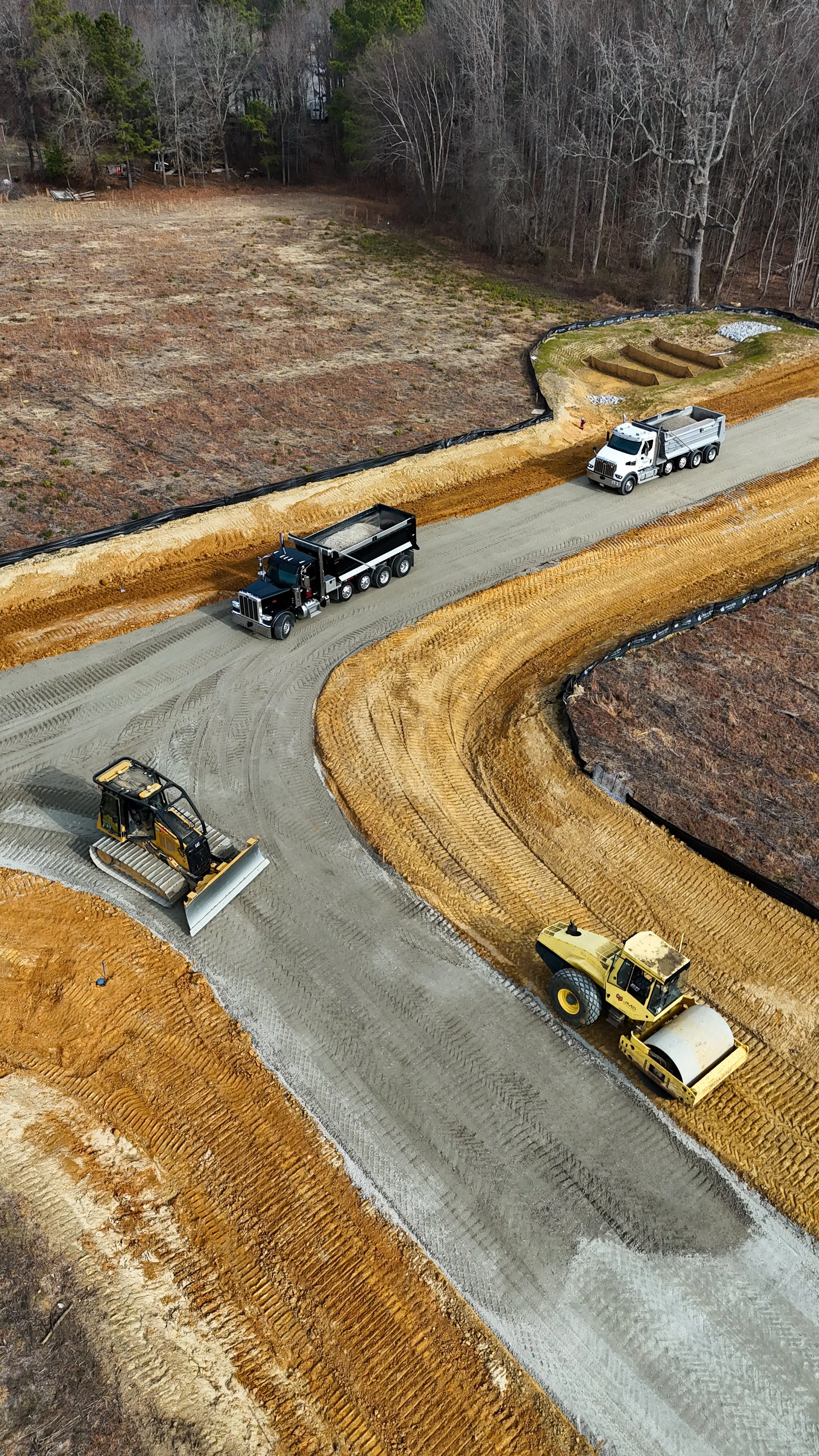 Aerial view of road construction with heavy machinery leveling and paving the road on a dirt path surrounded by trees.