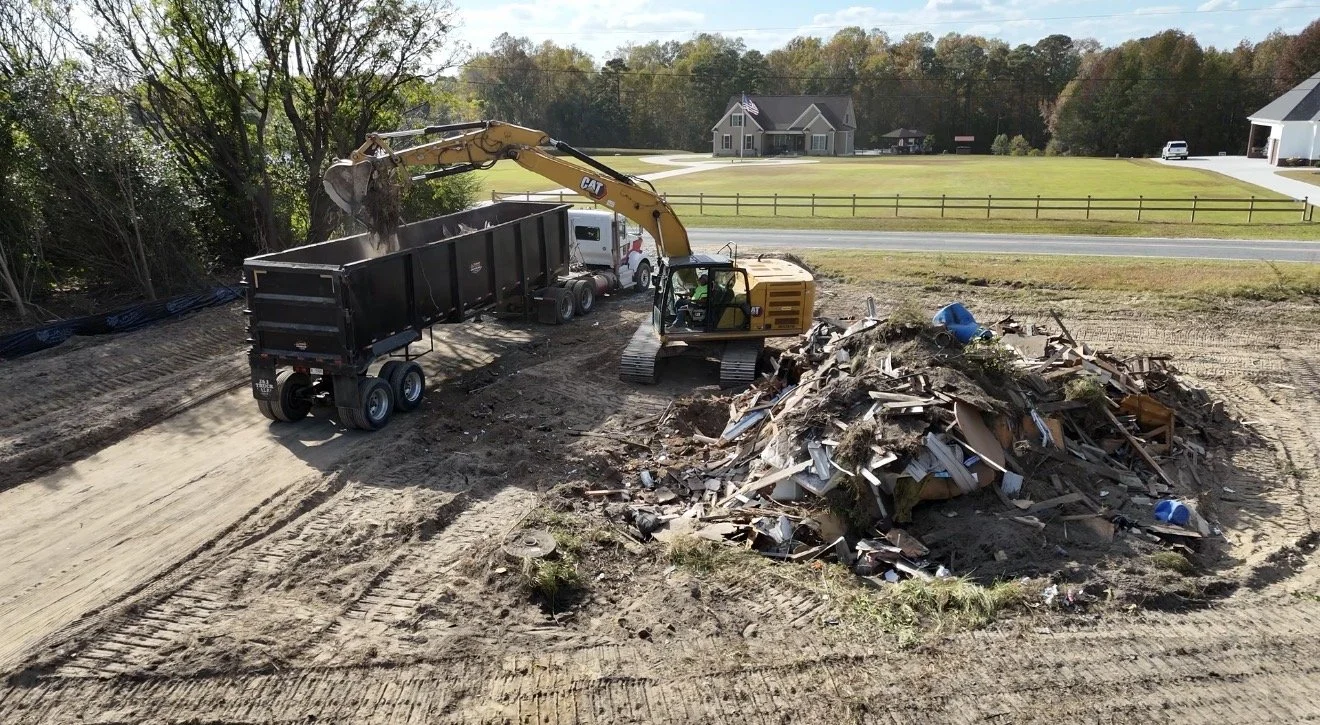 Construction site with excavator filling a large dump truck with debris, piles of rubble, and a suburban neighborhood in the background.