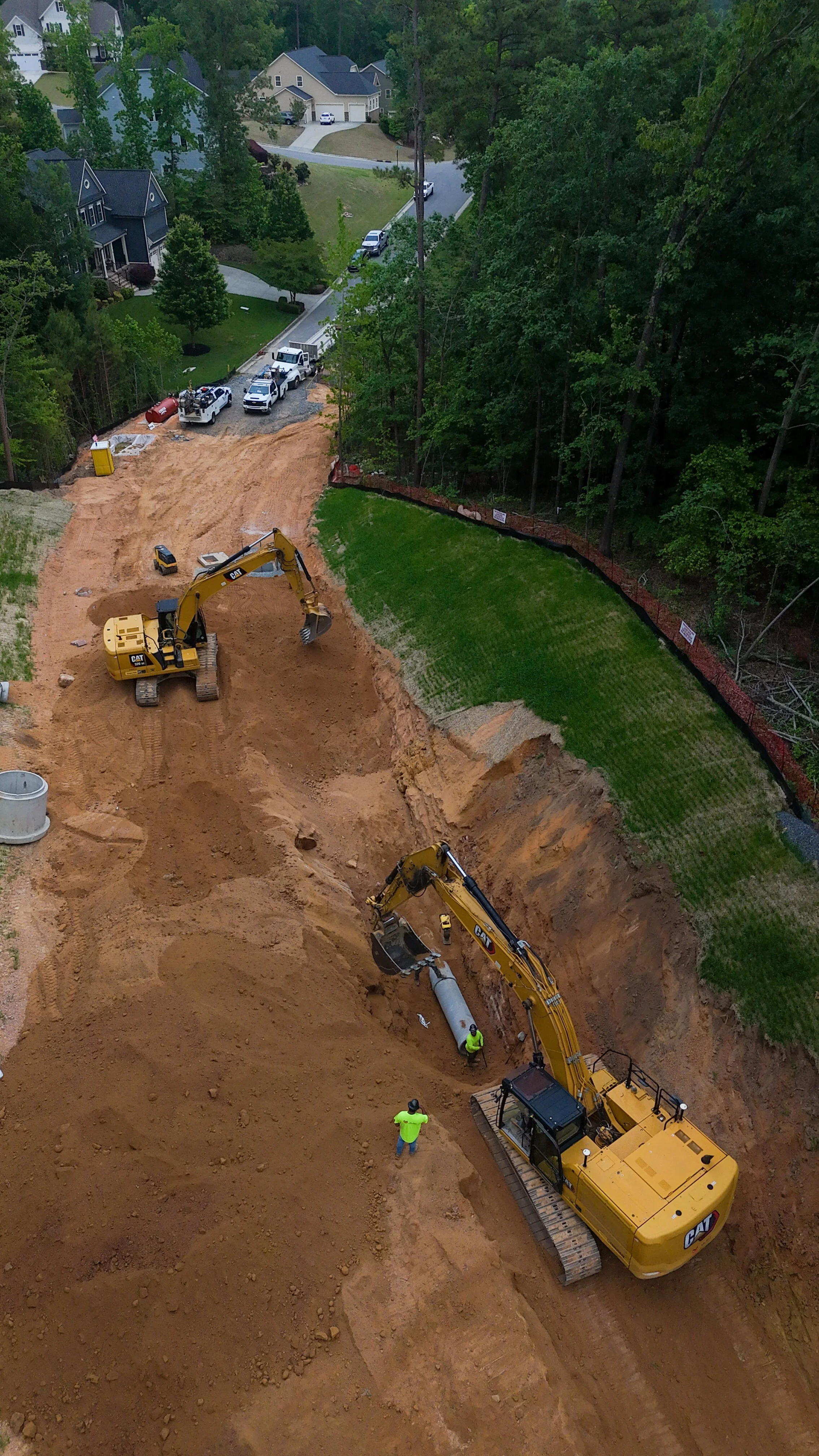 Construction workers and excavators working on a large earth excavation site next to a residential neighborhood with green lawns and trees.