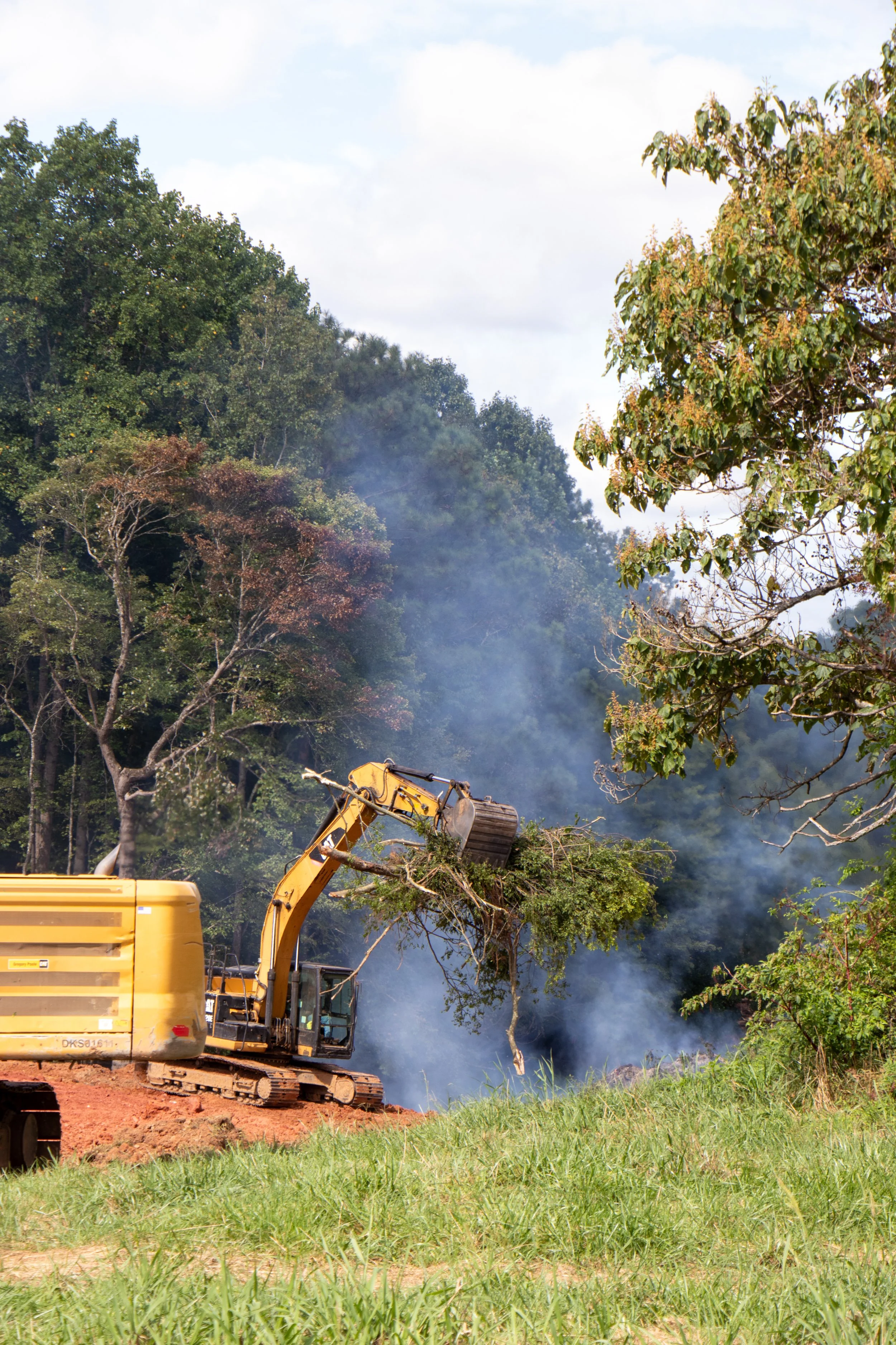 A yellow excavator tearing down trees in a green field with smoke rising in the background and a partly cloudy sky.