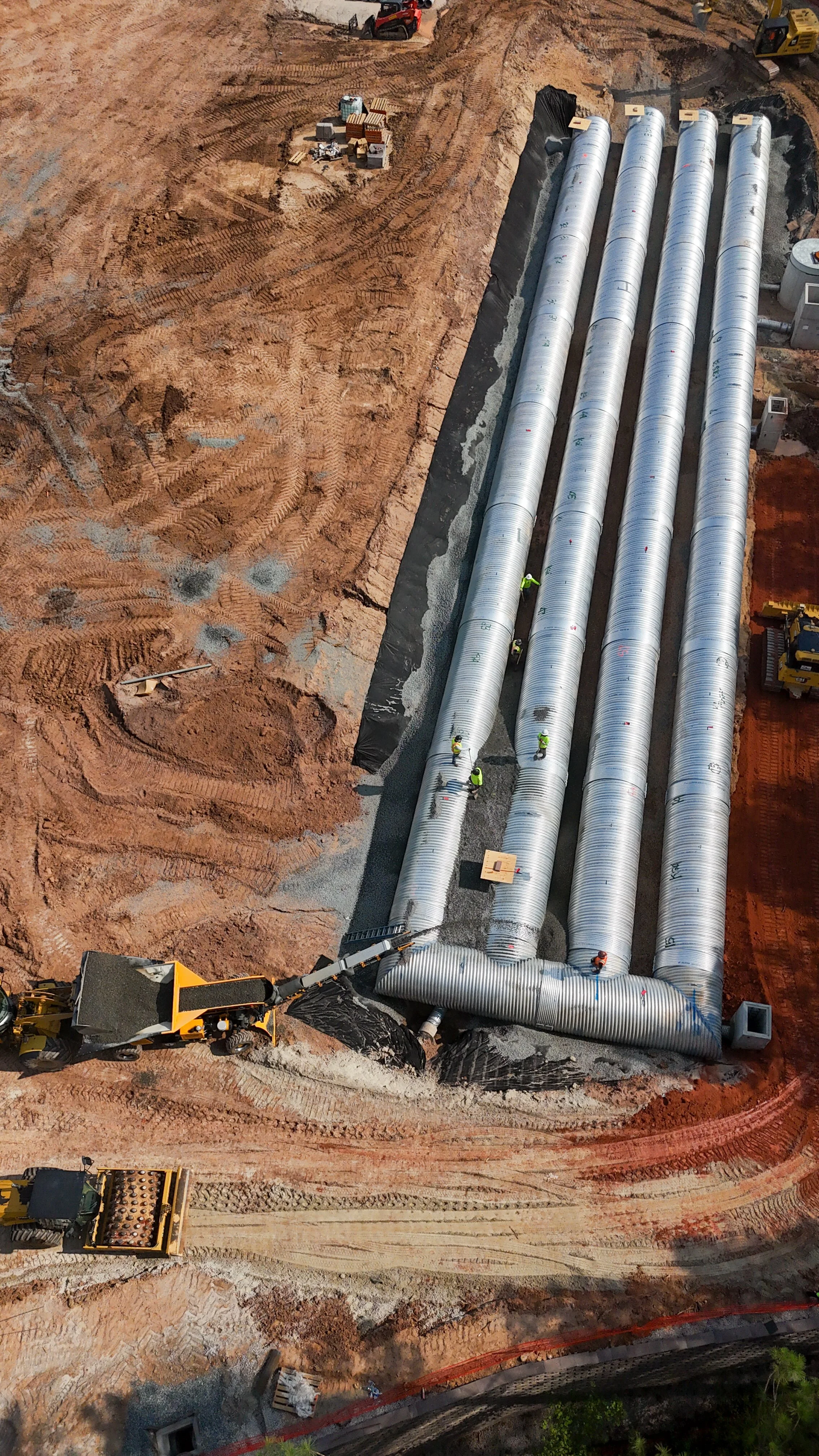 Construction site with large metallic pipes being installed underground, construction workers working, and earth-moving equipment operating on reddish dirt.