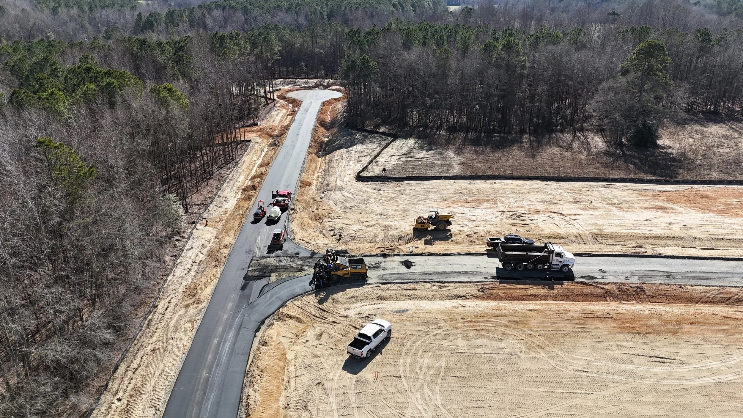 Aerial view of a road construction site with workers, heavy machinery, and vehicles working on paving a new road through a wooded area.
