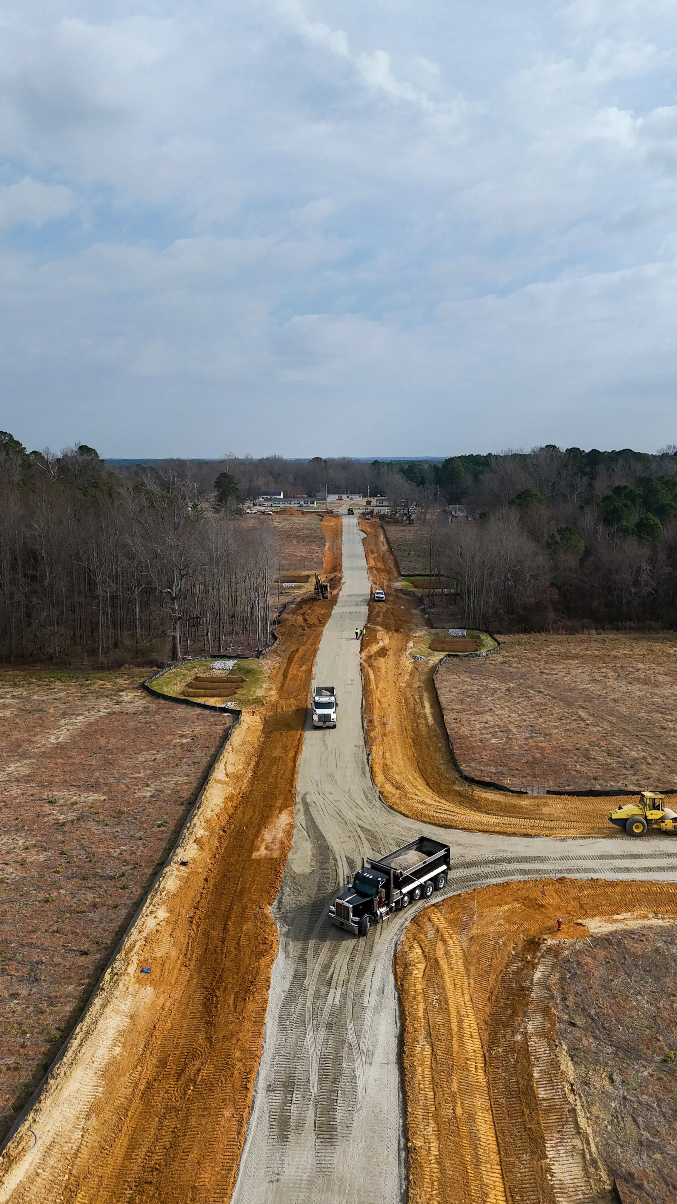 Construction workers and trucks working on a road construction site with a partially paved road surrounded by dirt, with a wooded area on the sides, under a cloudy sky.