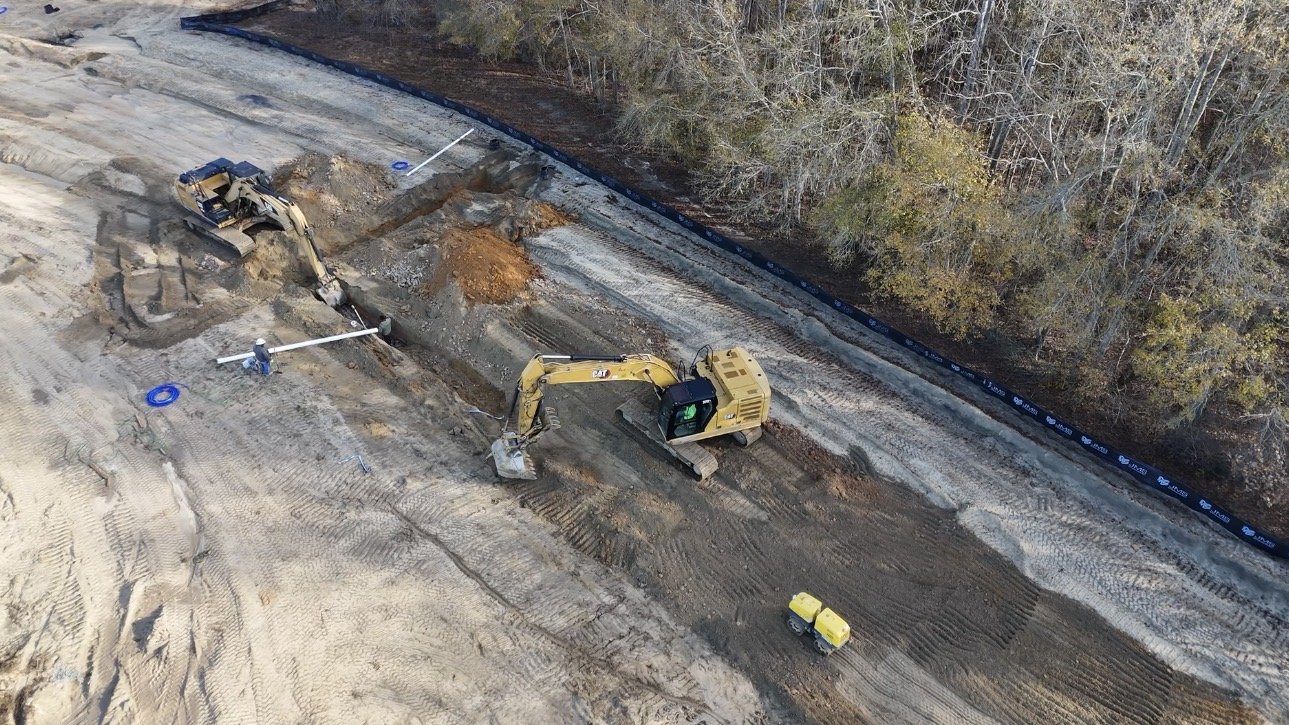 Construction site with excavators digging up dirt along a cleared area next to a tree-lined hillside.