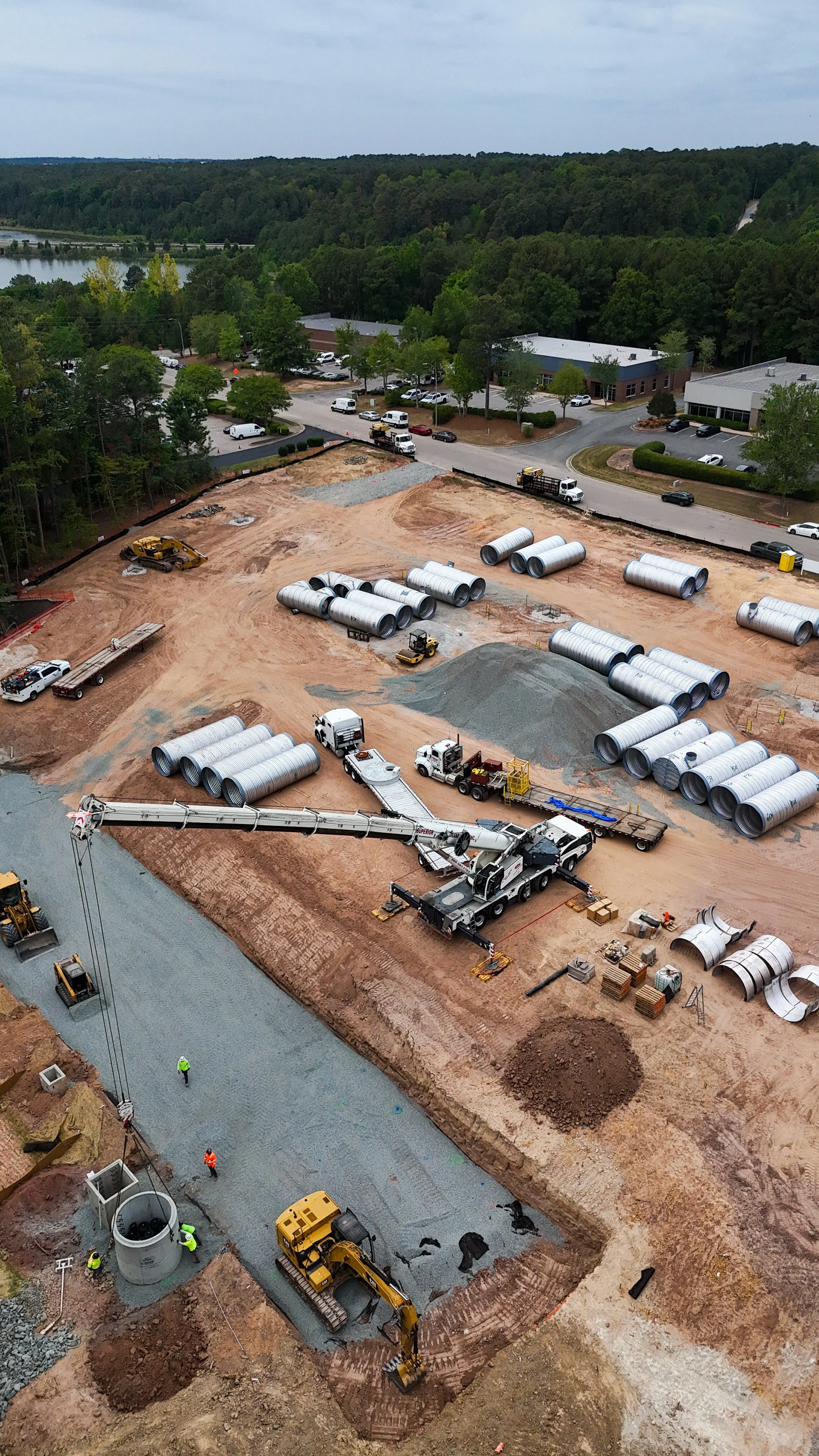 Construction site with large pipes, construction vehicles, and workers, with a background of parked cars, buildings, trees, and a river.