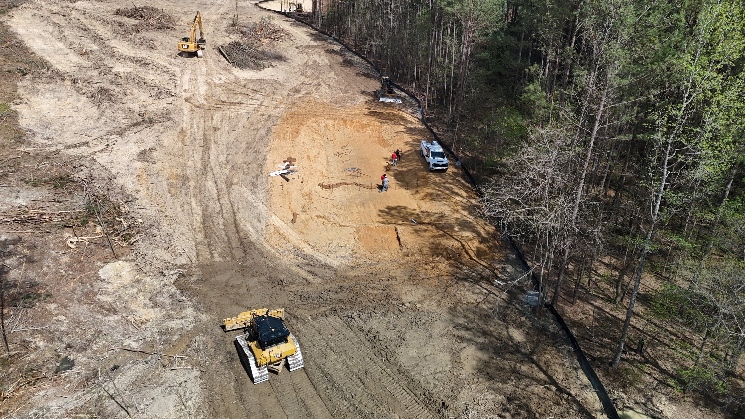 An aerial view of a construction site in a forested area, where workers are leveling the ground with machinery, including a bulldozer and an excavator, preparing the land for development.