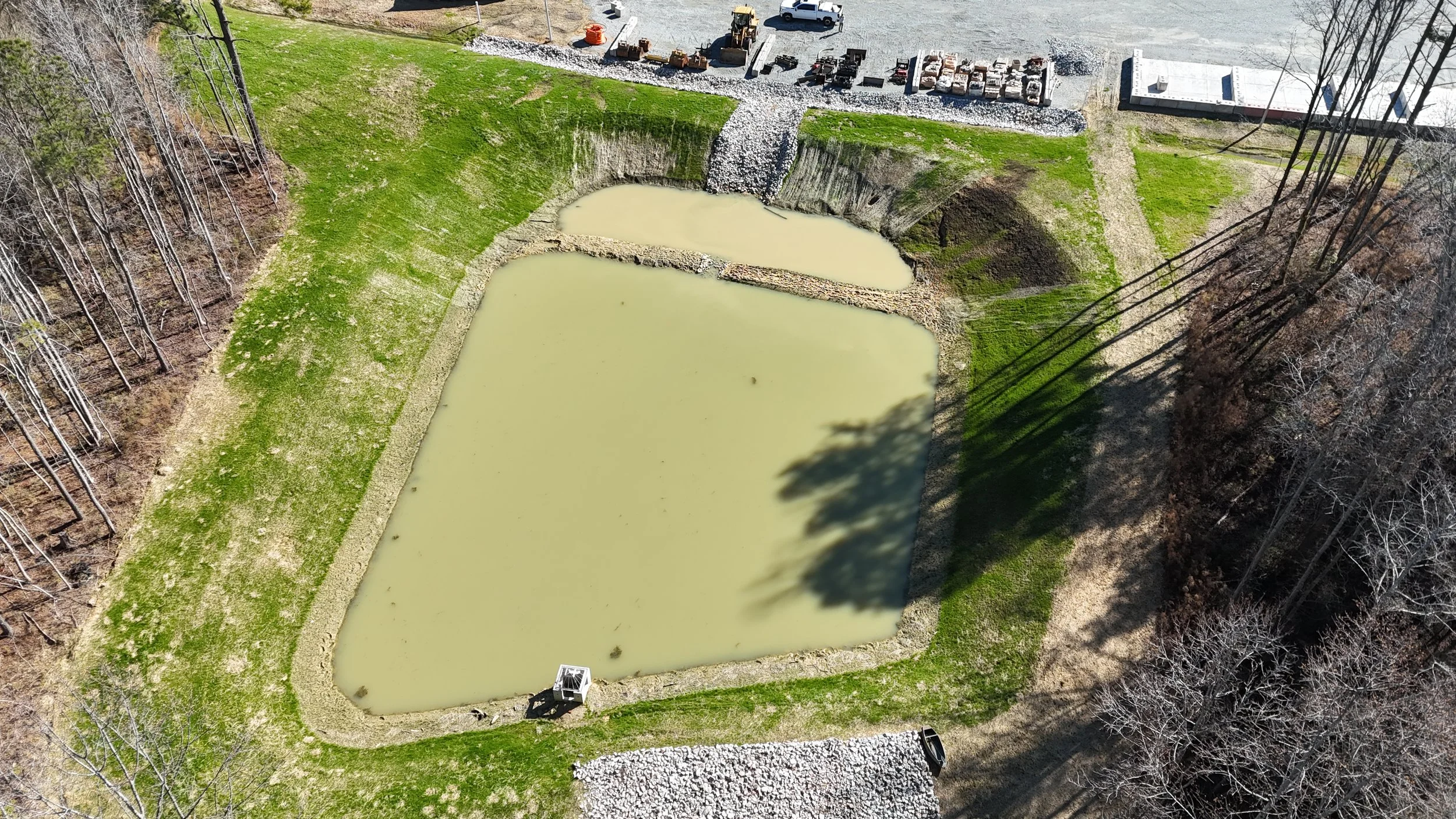 A flooded area around a small reservoir or pond, with murky green water, surrounded by grass and trees, and construction equipment and vehicles at the top of the image.