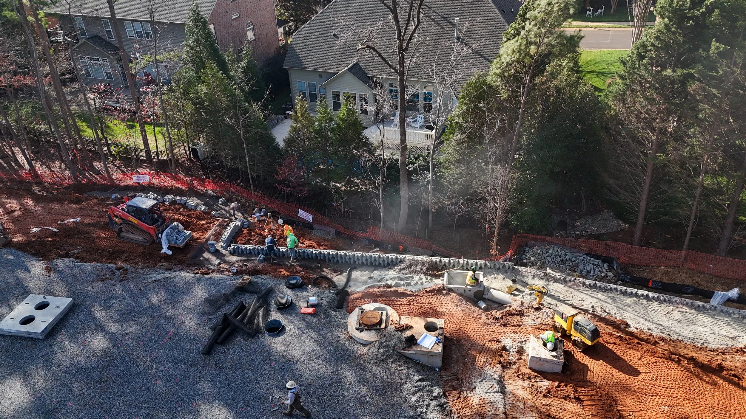 Construction workers building a road or driveway with orange dirt, black pipes, and concrete blocks, next to residential houses with trees and a street in the background.