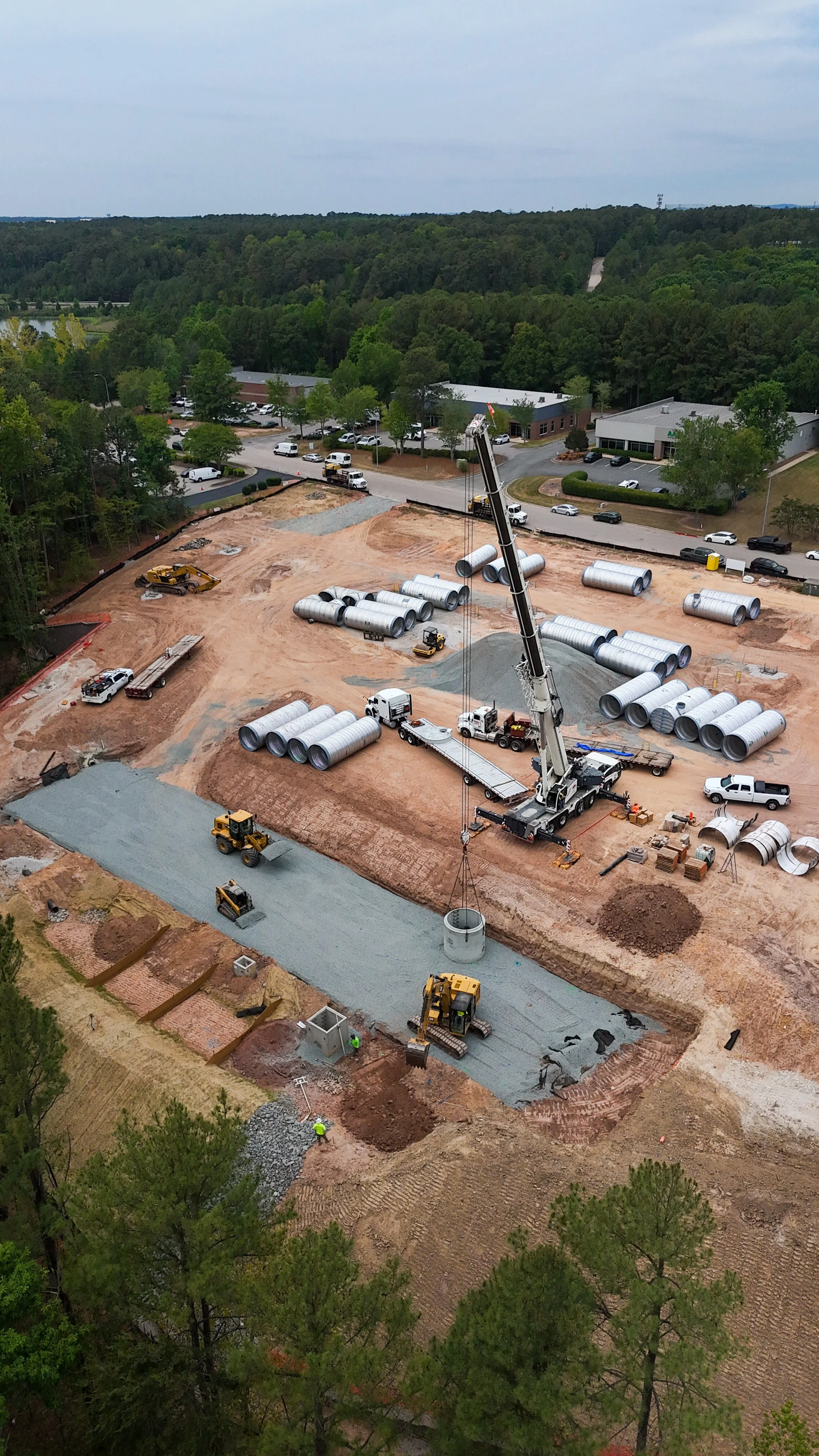 Construction site with large pipes, cranes, trucks, and workers on a project to develop a building or infrastructure, surrounded by trees and nearby roads.