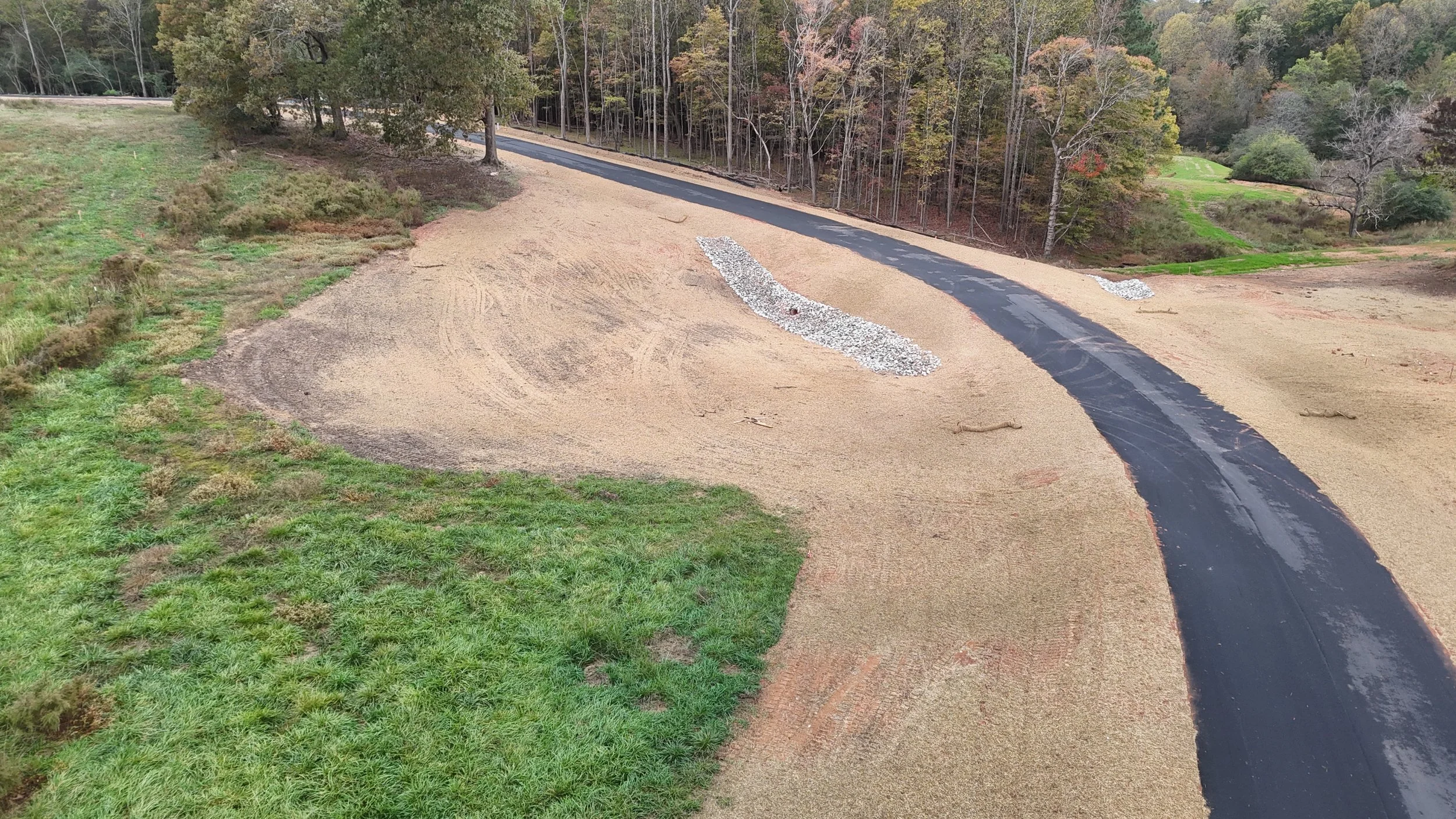A golf course undergoing renovations with patches of sand and gravel, a paved path, and surrounding trees with autumn foliage.