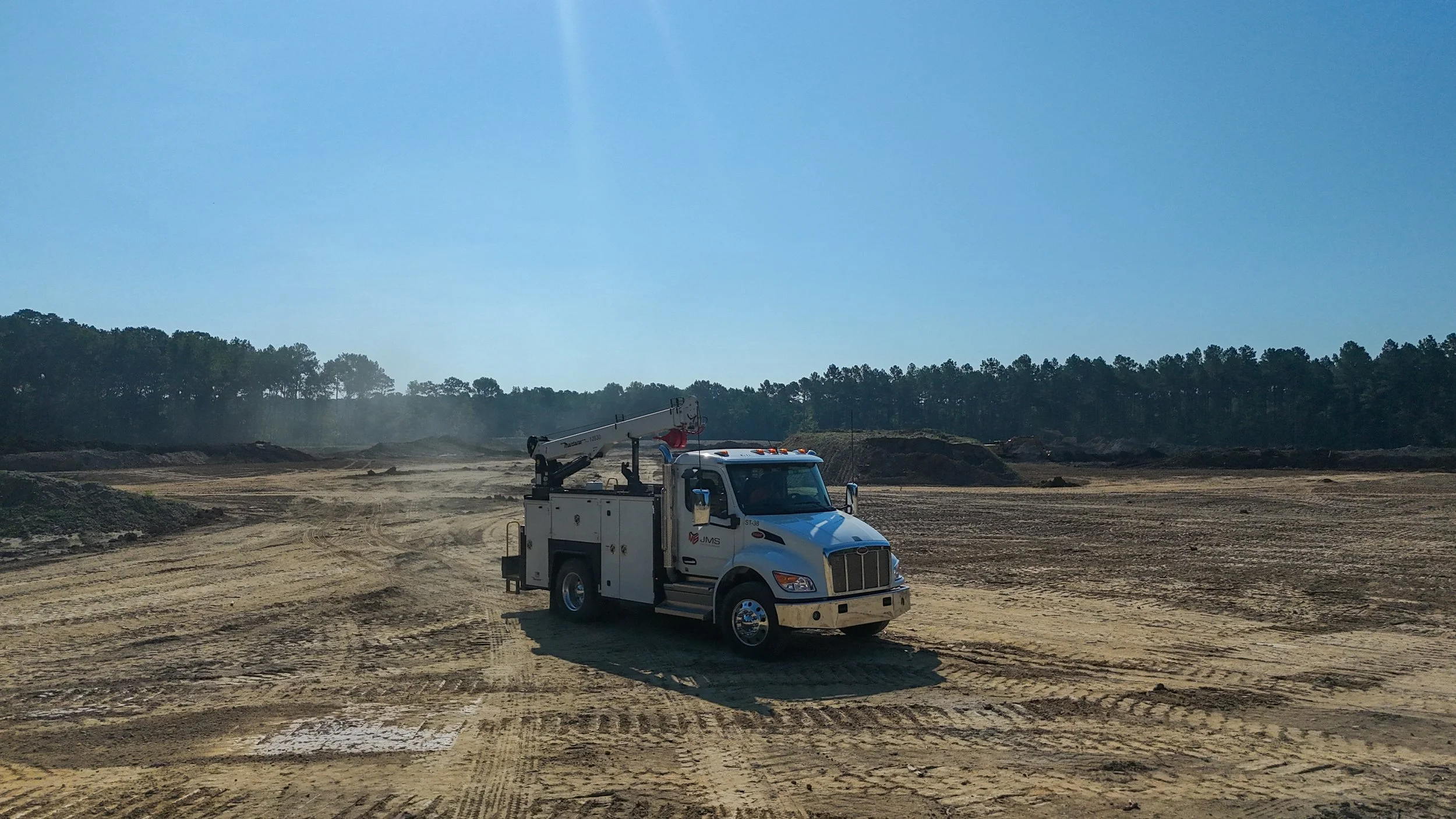 Construction site with a white utility truck, dirt ground, and a forest in the background under a clear blue sky.