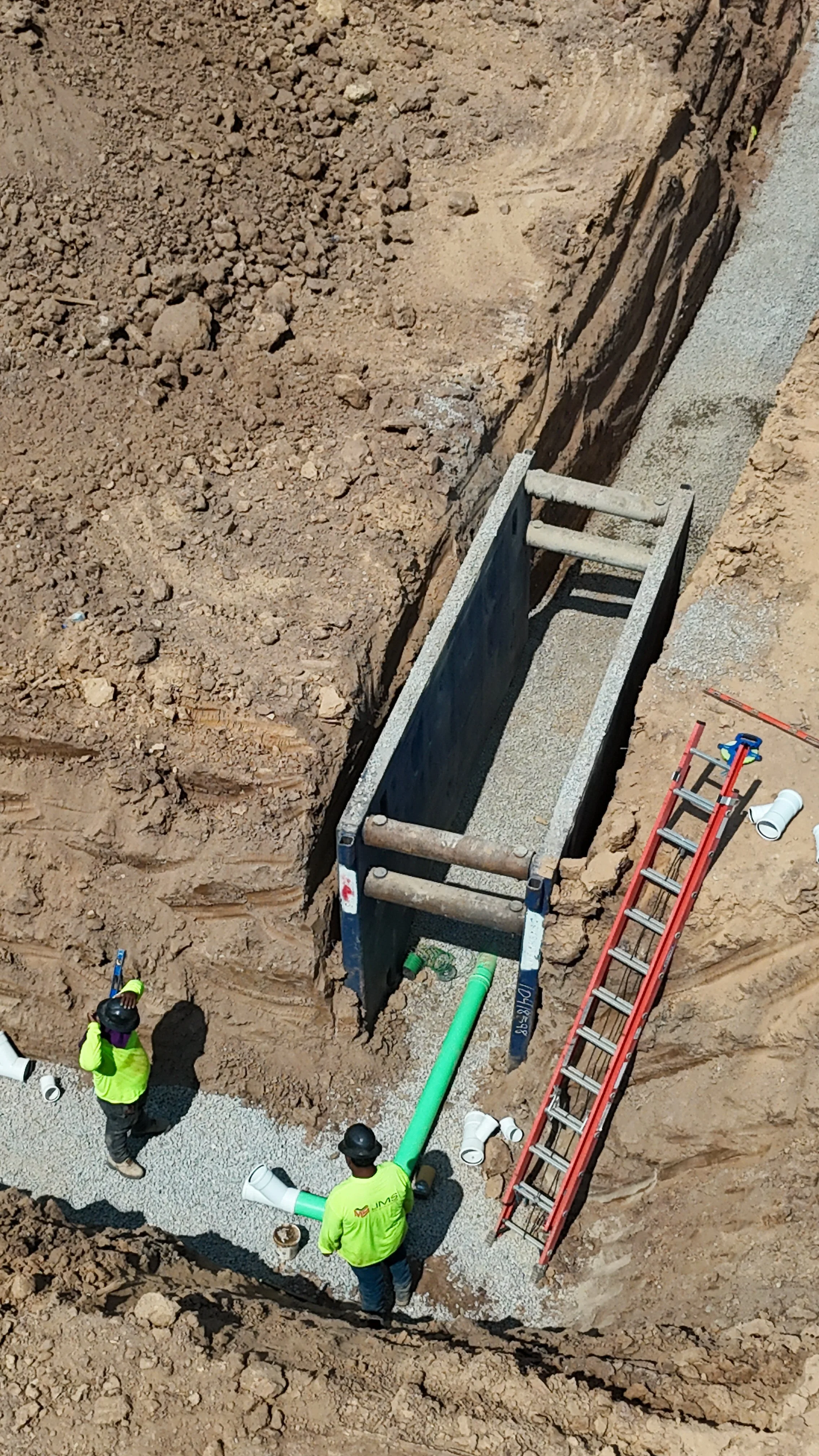 Construction workers installing underground piping in a trench with gravel foundation, using ladders and pipes.