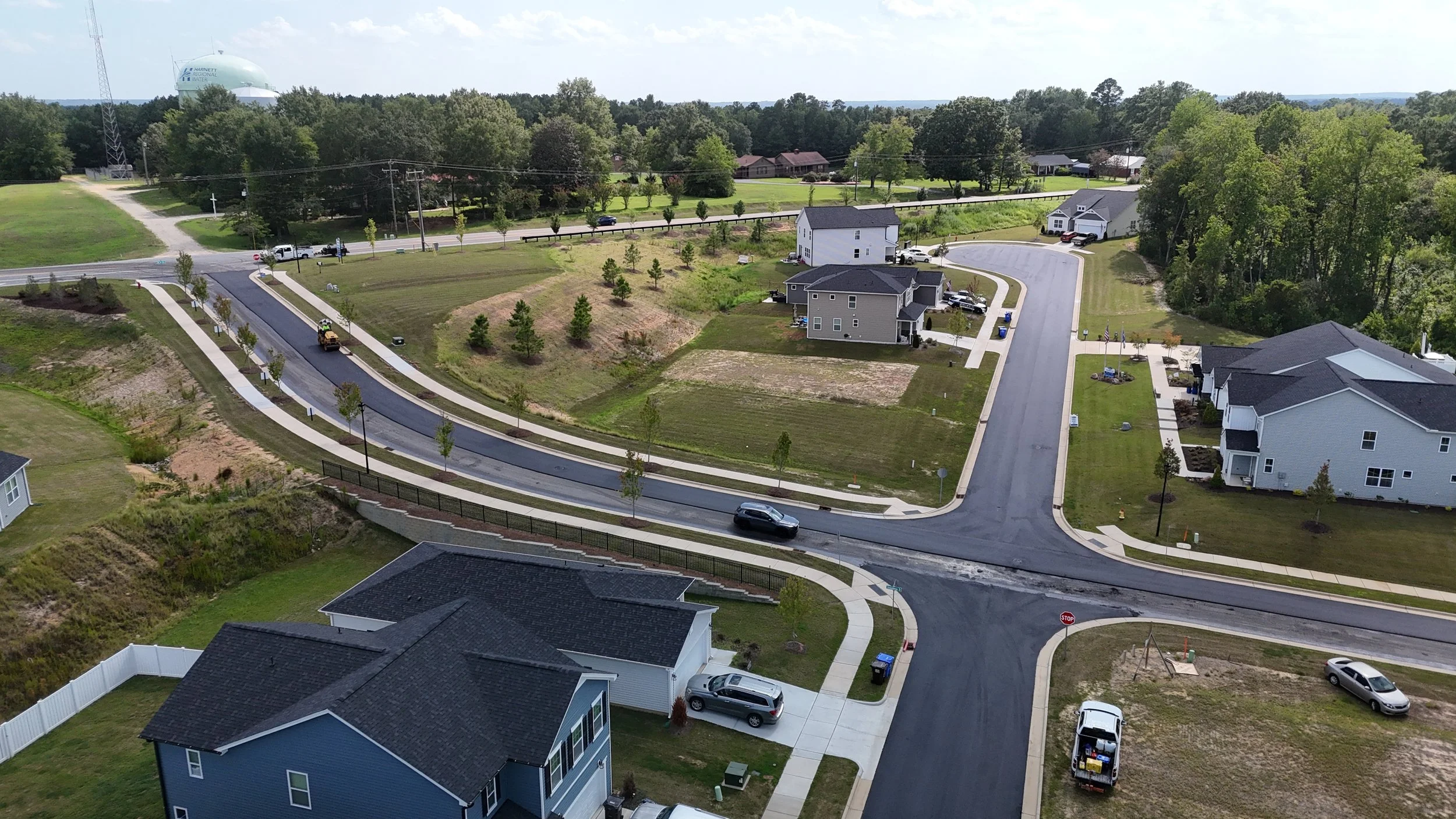 Aerial view of a suburban neighborhood with newly paved roads, several houses with driveways, green lawns, and trees. A construction vehicle is working on a curved road, and traffic is light with a few cars parked and driving.