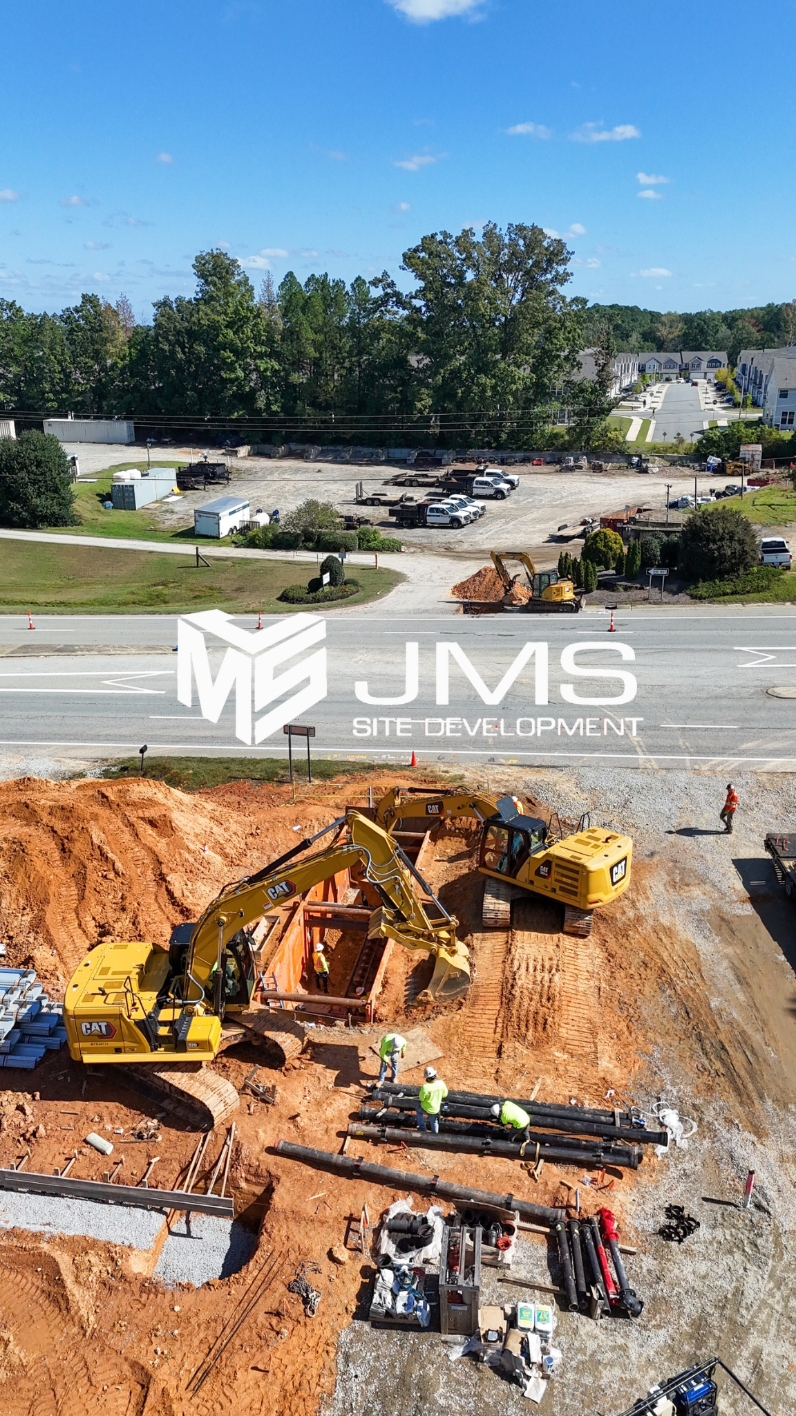 Construction site with excavators digging and installing pipes, workers in safety gear, parking lot with cars and trees in the background.