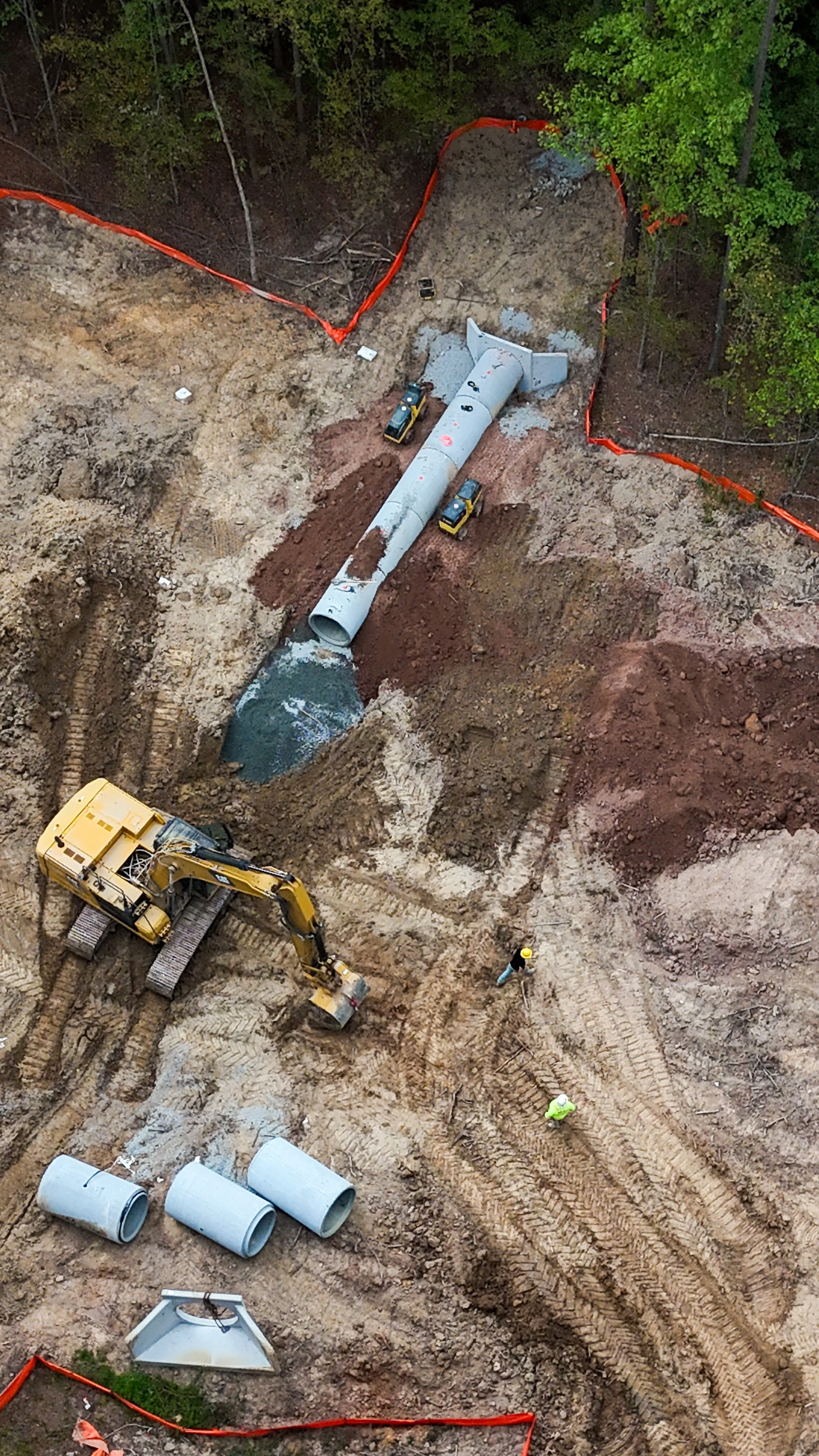 Construction site with large vertical pipeline, construction vehicles, workers, and piping materials, surrounded by dirt and trees.