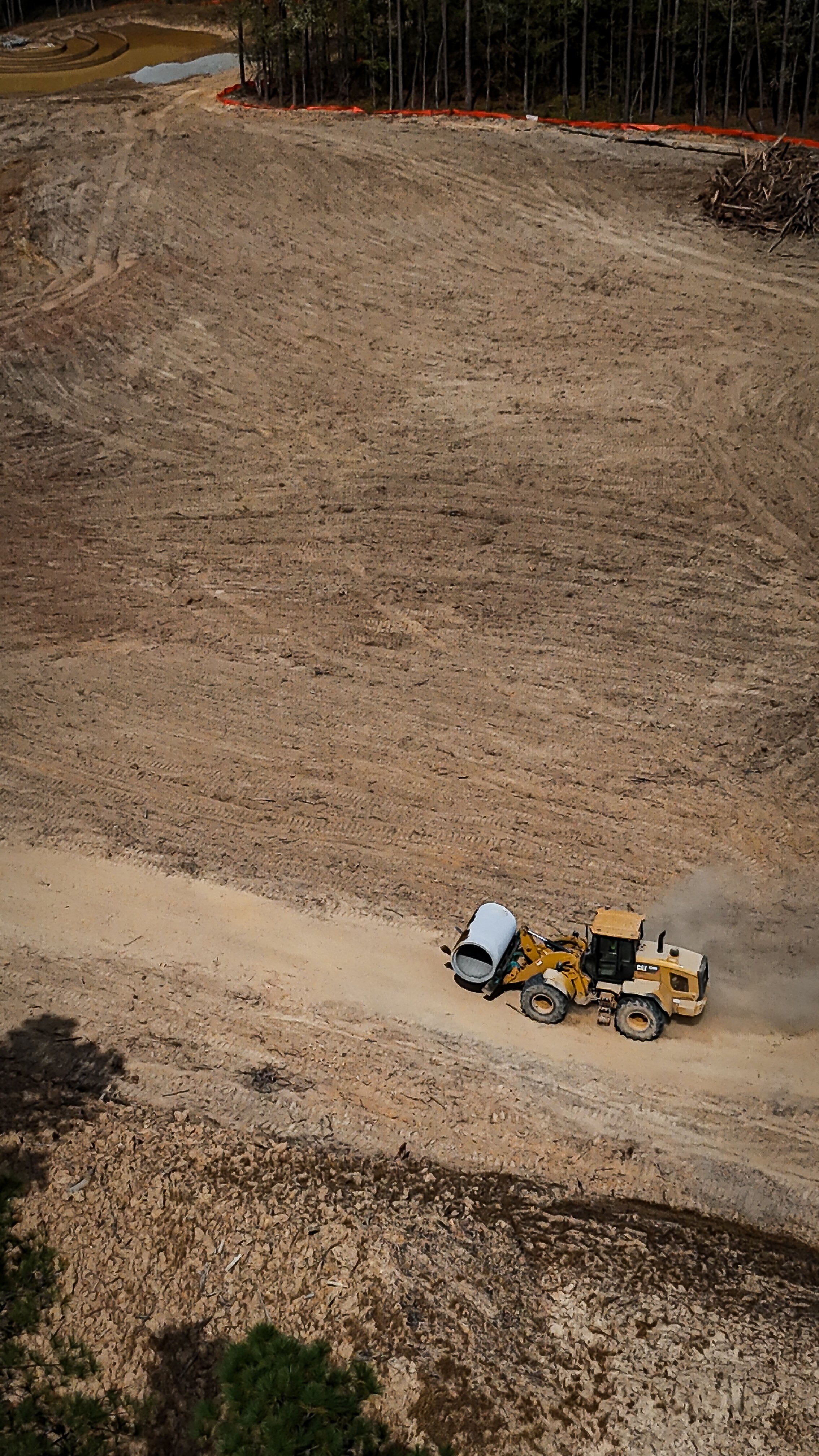 Construction site with a yellow cement mixer truck and a concrete mixer. Dirt and earth-moving equipment in a large, partly cleared area with trees in the background.
